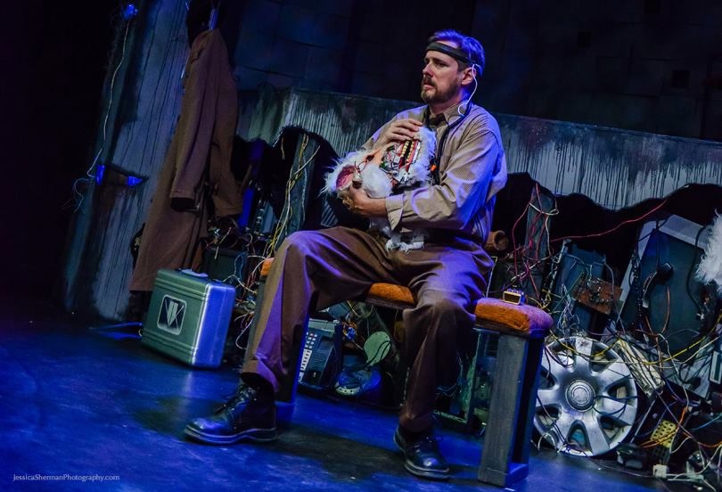 Man sitting on a chair holding a microphone, stage with props and equipment in background.