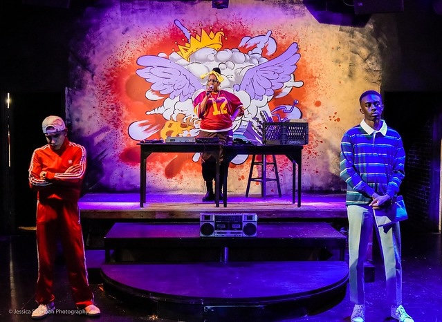 Three people on stage with a colorful angel mural background, one person at a DJ booth, two others standing on stage.