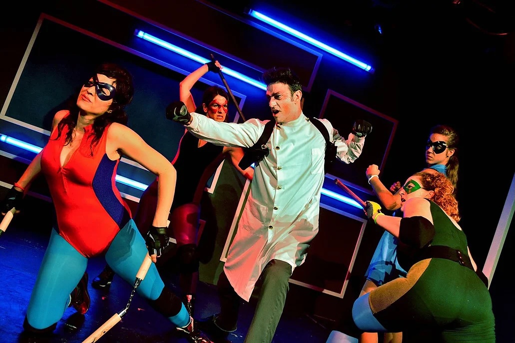 Group of five people in colorful costumes dancing in a room with neon lights, some wearing sunglasses and gloves.
