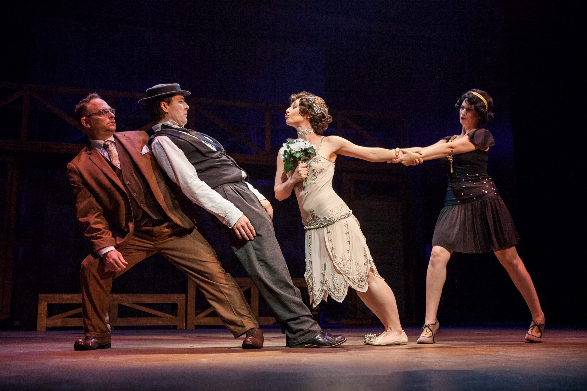Four performers on stage, two men and two women, engaged in a dance or theatrical scene, with dark background and stage lighting.