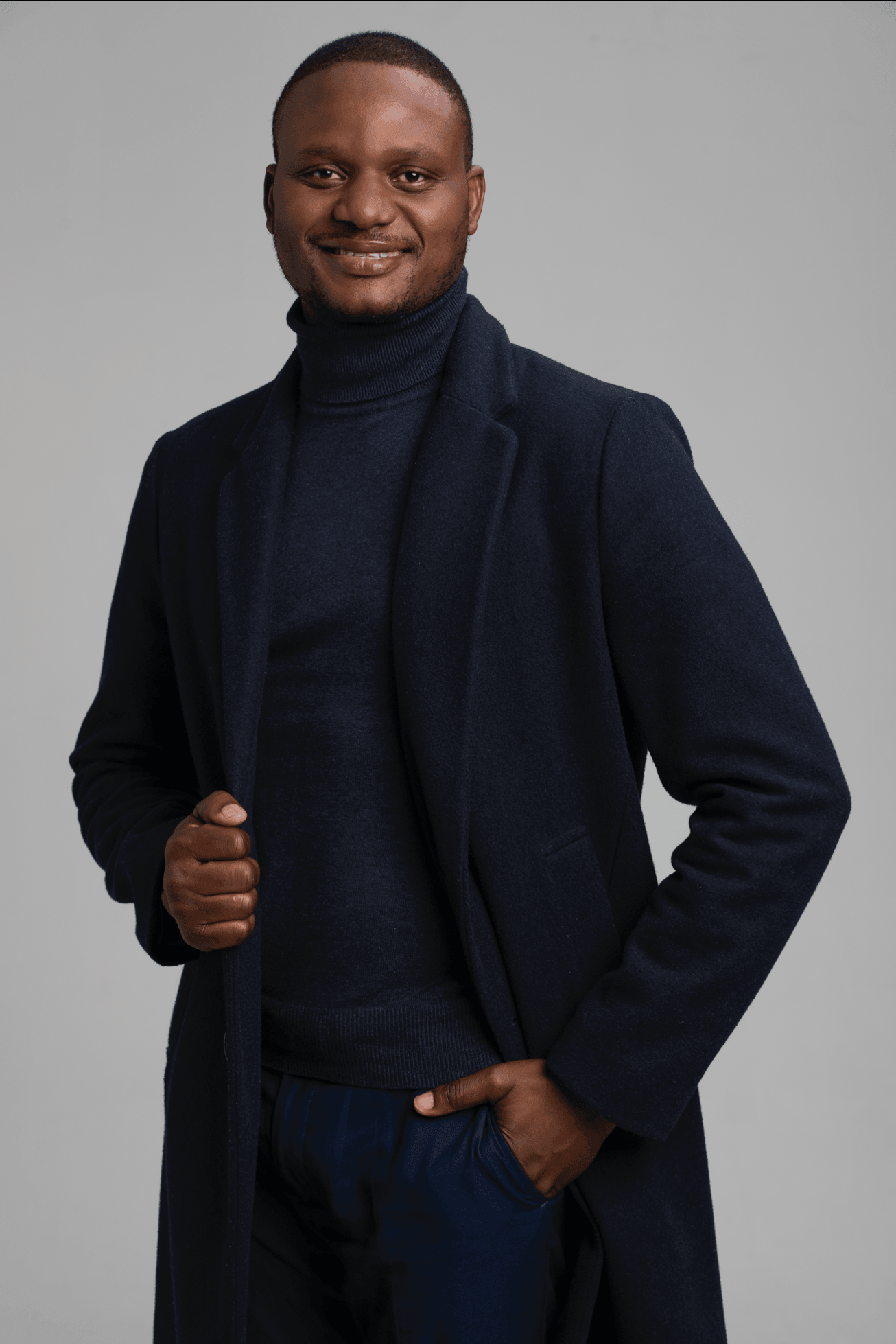 Man in dark coat and turtleneck smiling, standing with one hand in pocket against a plain background.