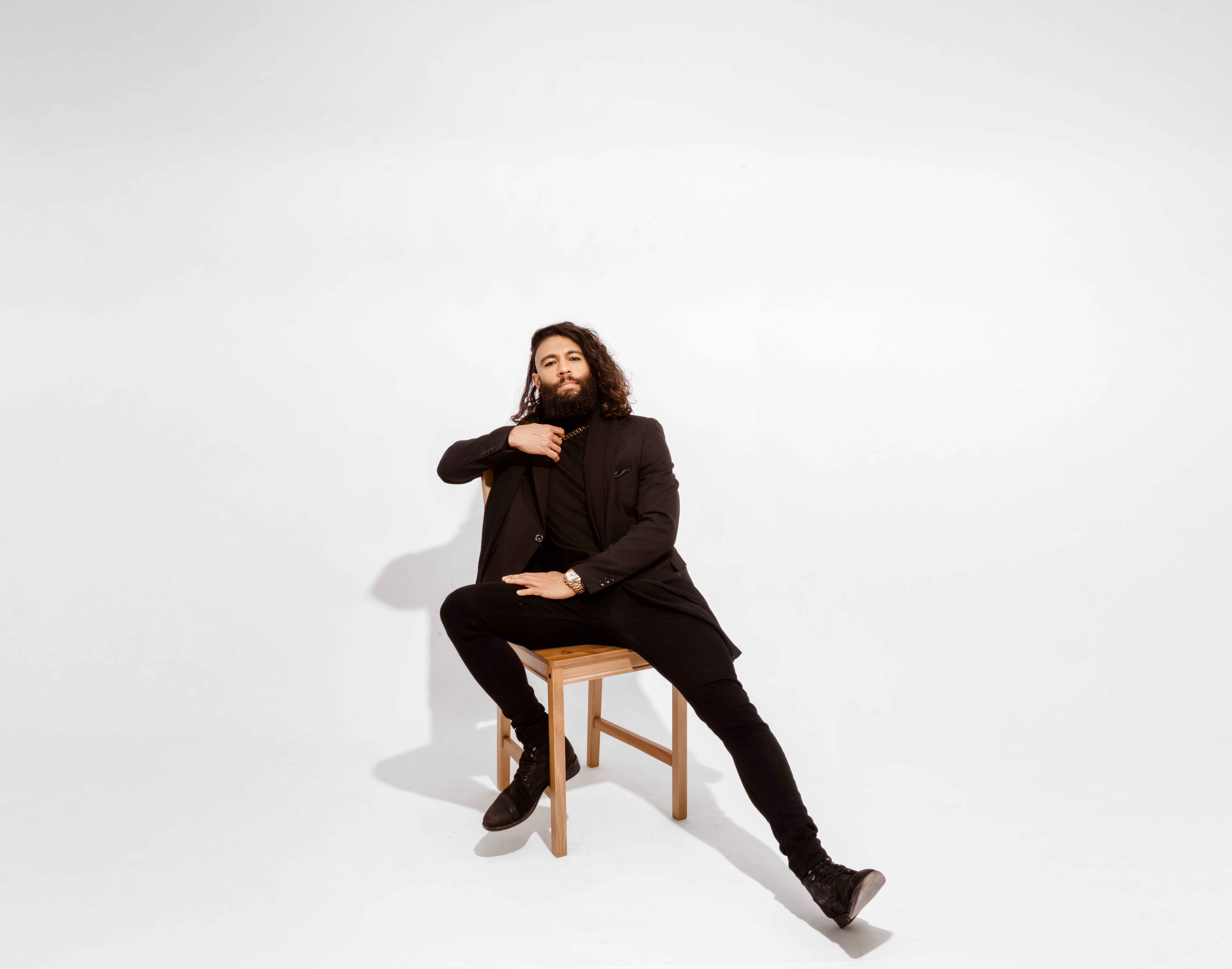 Person with long hair and beard sitting on a wooden chair, wearing dark clothing, against a plain white background.
