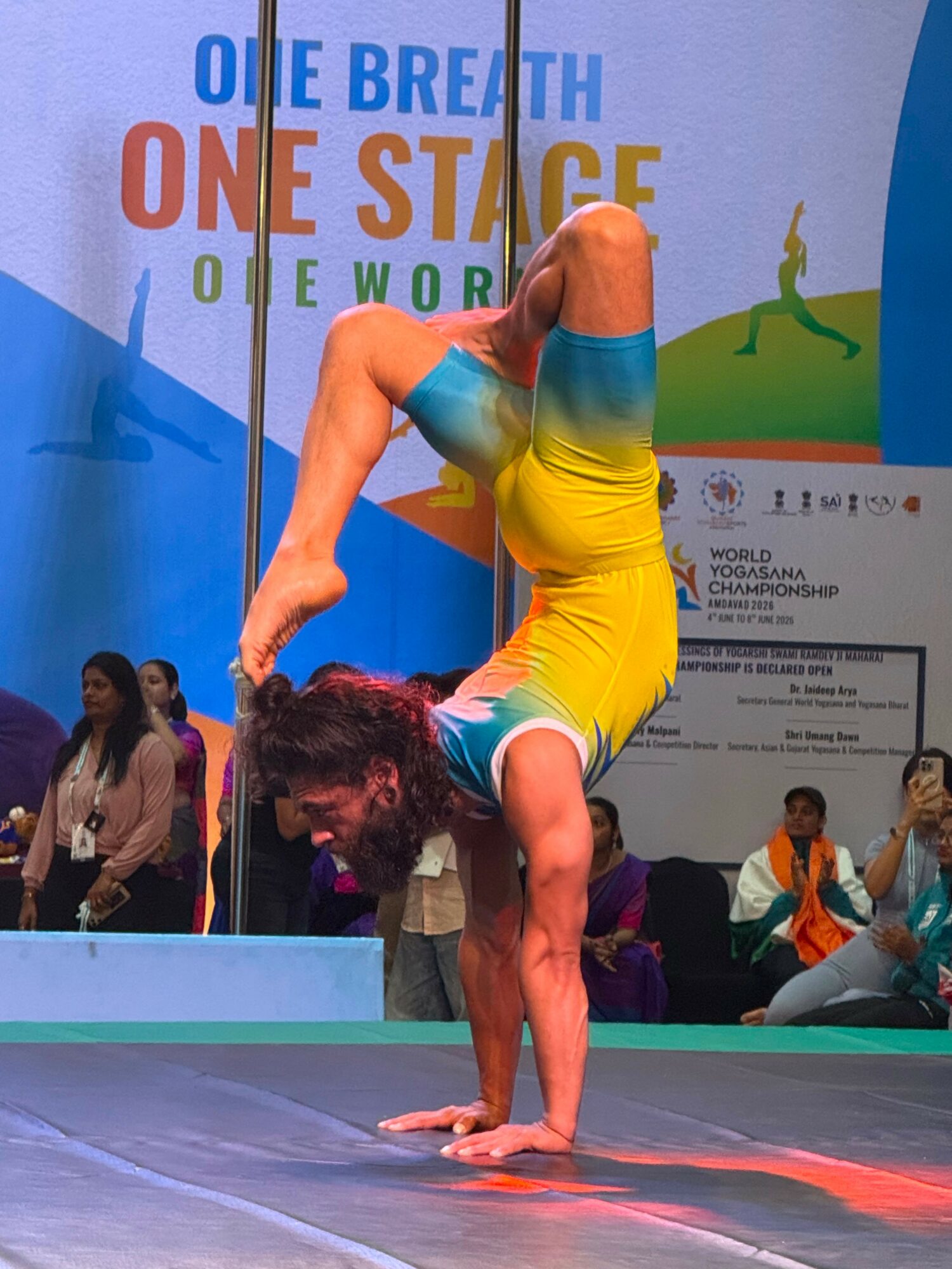 A person performs a handstand on a stage, wearing a yellow and blue outfit, with a colorful background and audience.