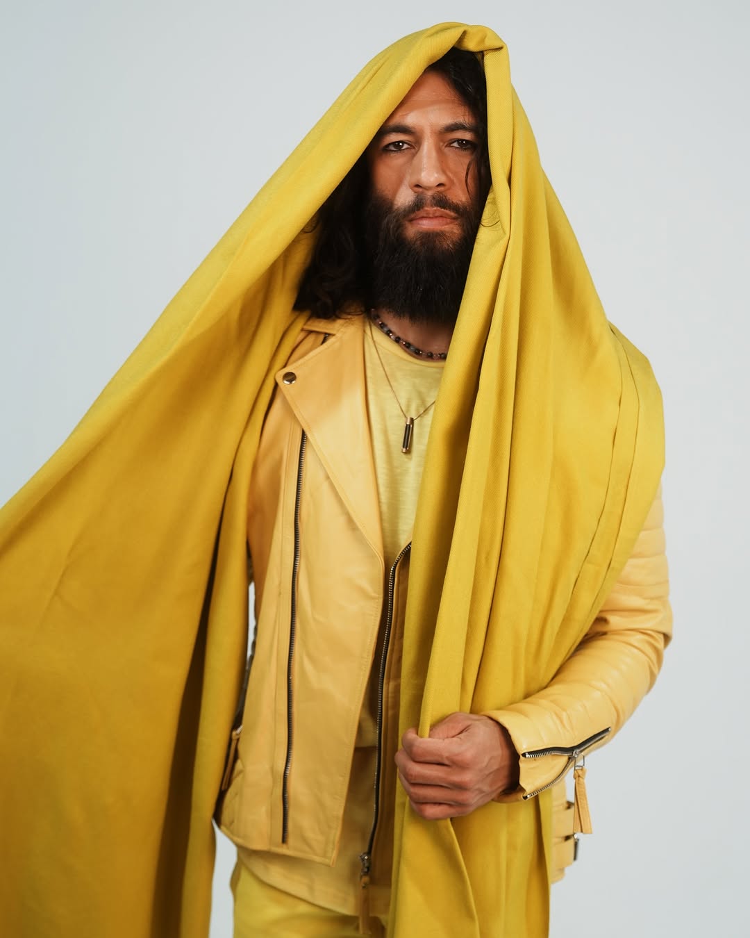 Man with long hair and beard wearing a yellow jacket and a yellow cloth draped over his head, standing against a plain background.