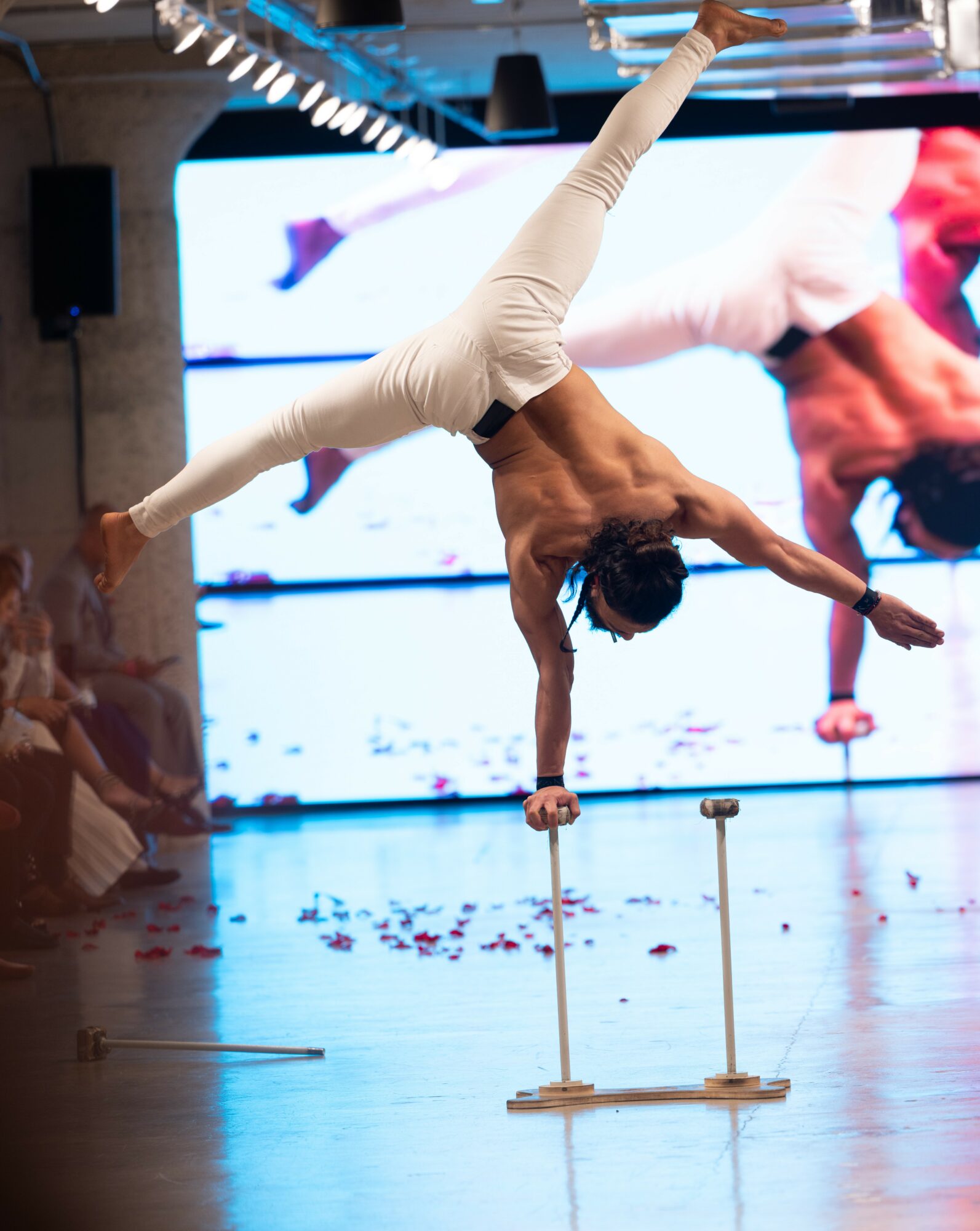 Performer balancing on hand with legs extended sideways, on two small platforms, in front of a large screen, audience watching.