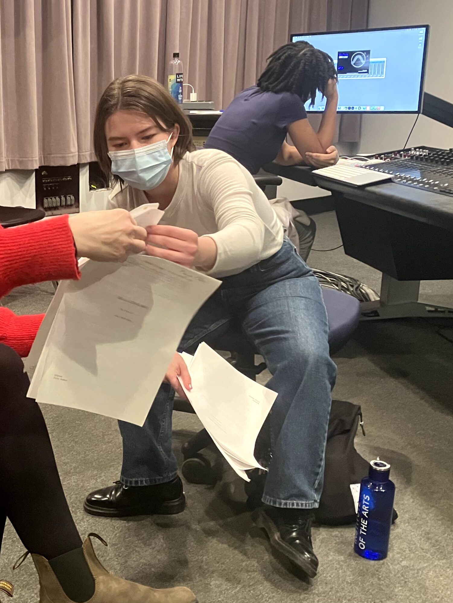 Person in a white shirt and face mask receives papers from another person, with a person working on a computer in the background.