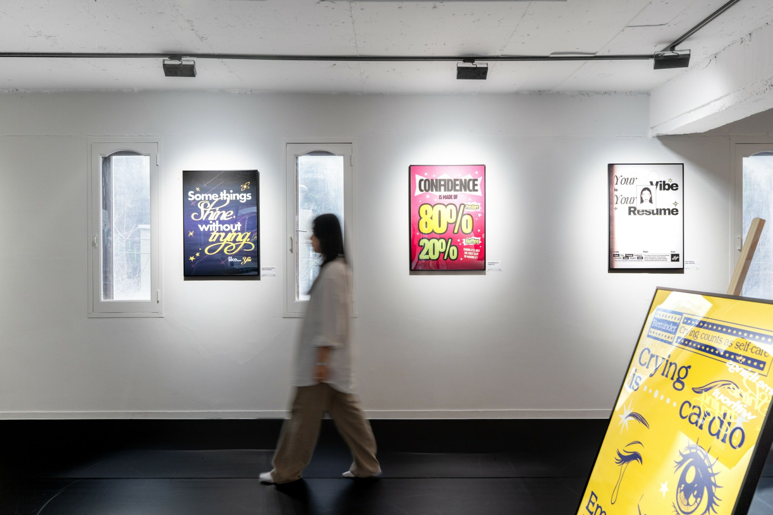 Person walking past a white gallery wall with four posters, one partially visible in the foreground, in an art gallery or exhibition space.