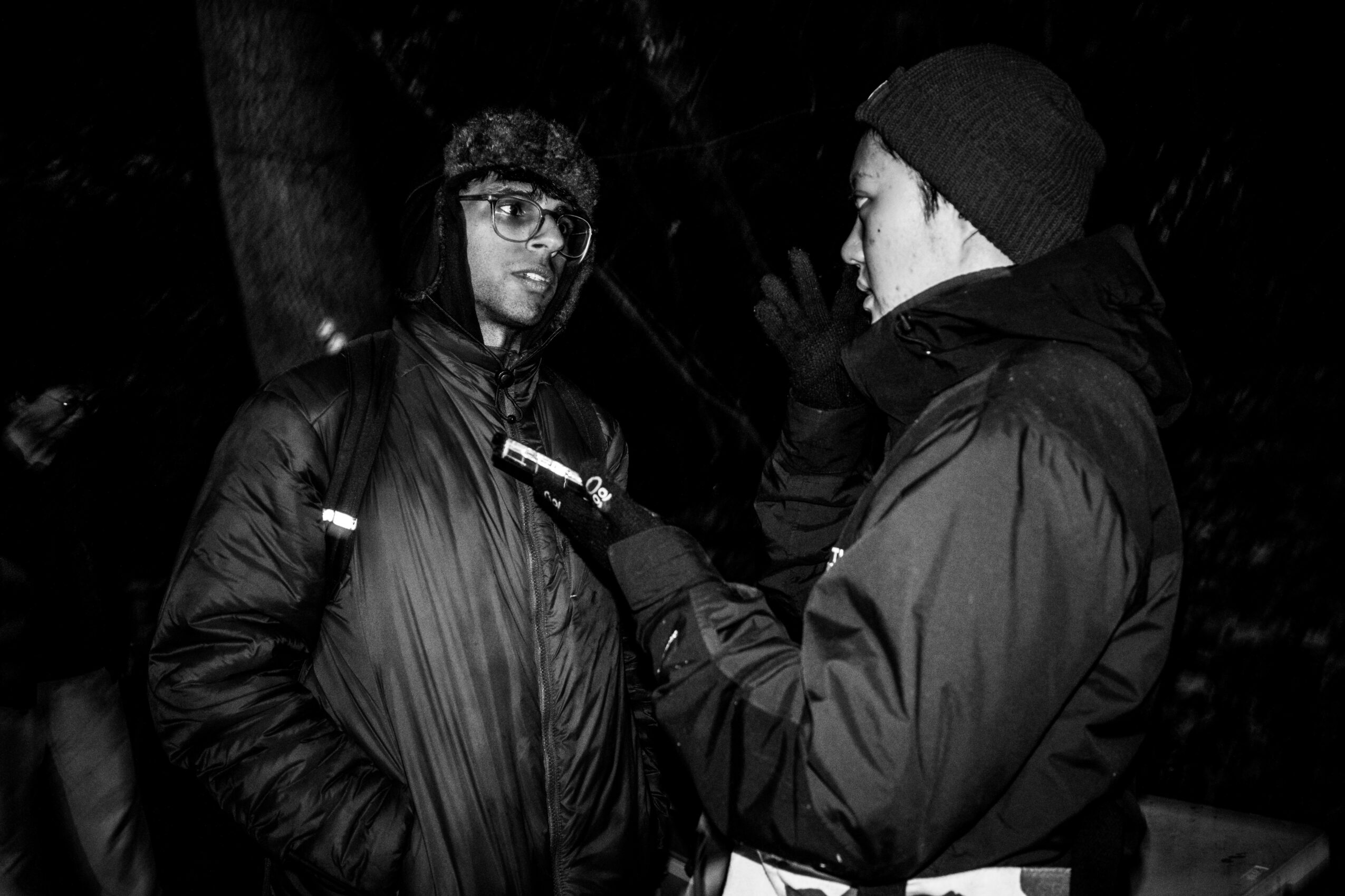 Two people in winter clothing talking outdoors at night, one holding a microphone, dark background.