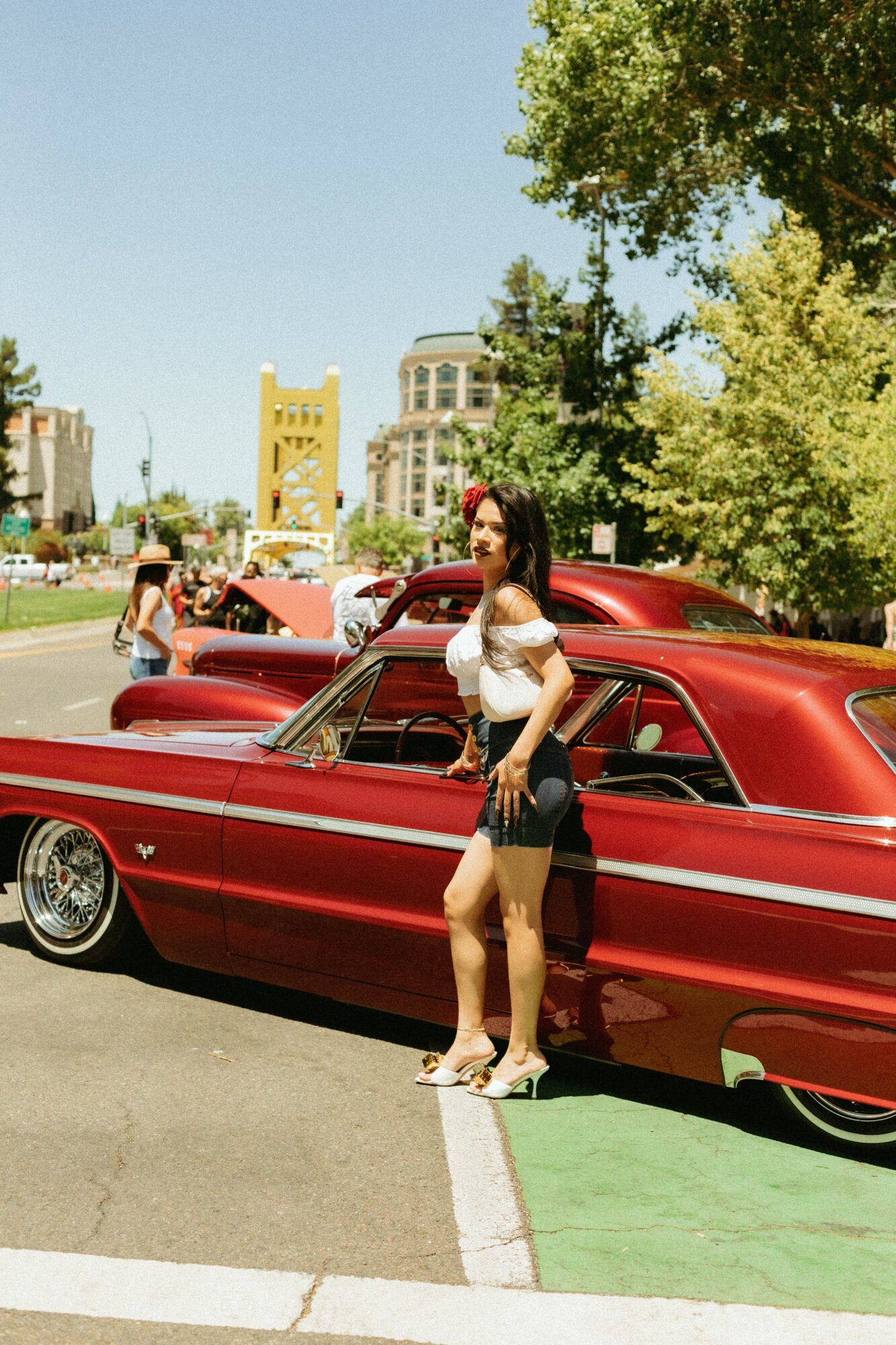 Woman standing next to a red vintage car outdoors on a sunny day, with trees and buildings in the background.