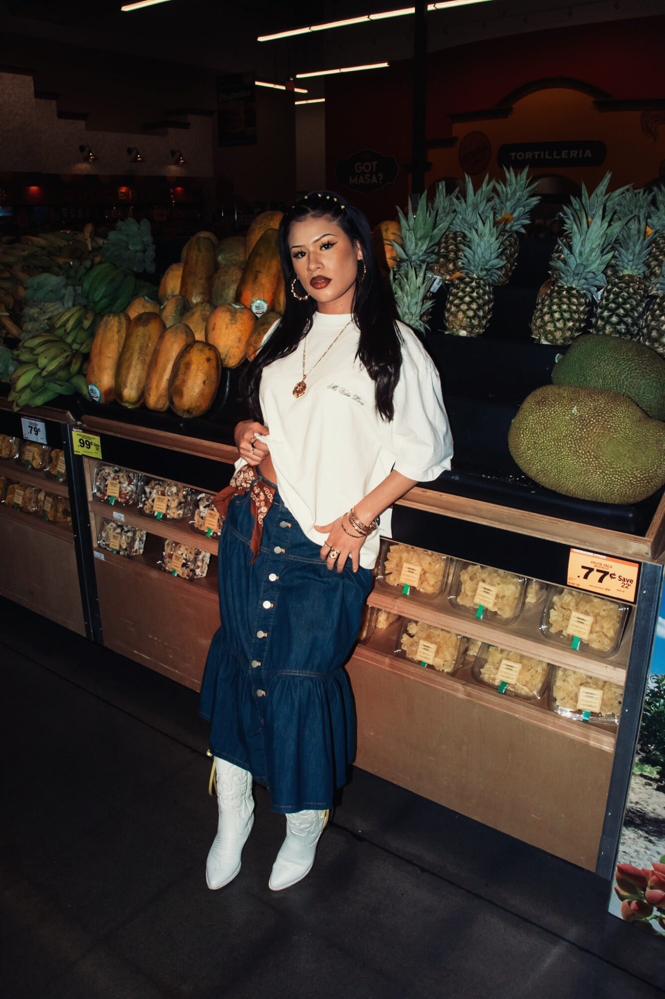 Woman with dark hair and red lipstick stands in a grocery store aisle with pineapples and melons behind her, wearing a white blouse and blue skirt, white boots.