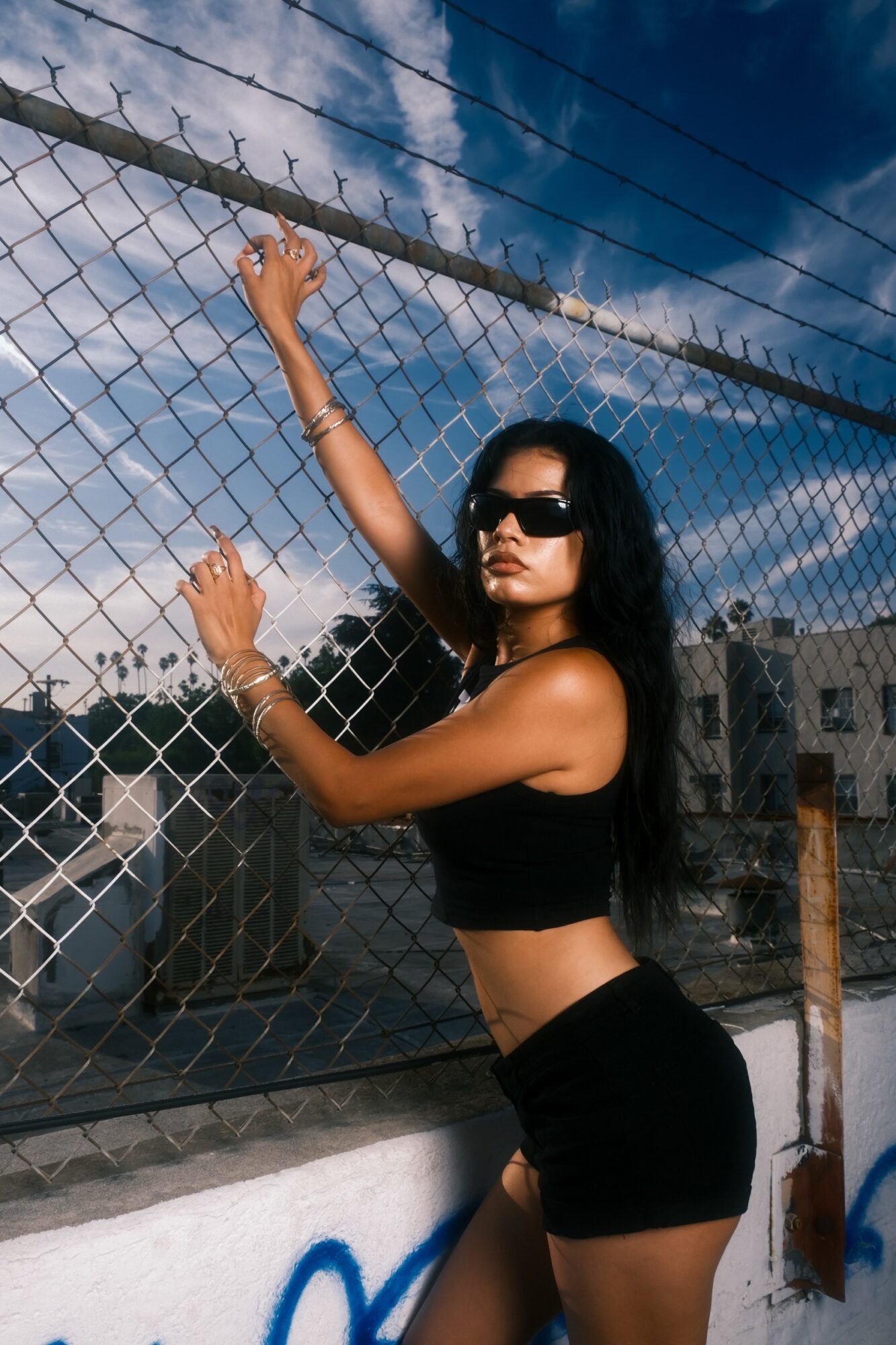 Woman with long dark hair and sunglasses stands by a chain-link fence, wearing a black crop top and shorts, against a blue sky.