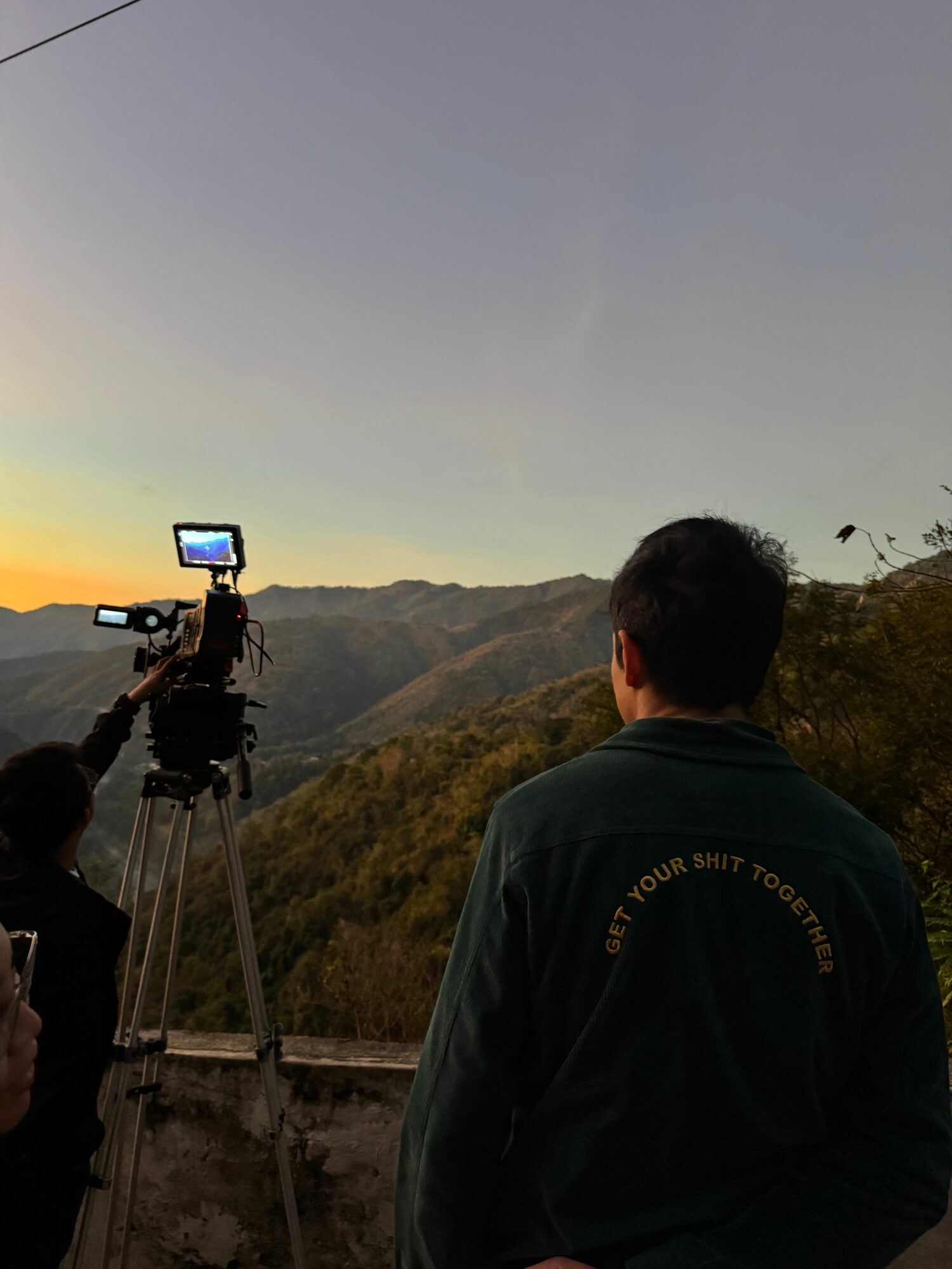 Person standing outdoors on a hill at sunset, with a camera on a tripod facing the landscape.