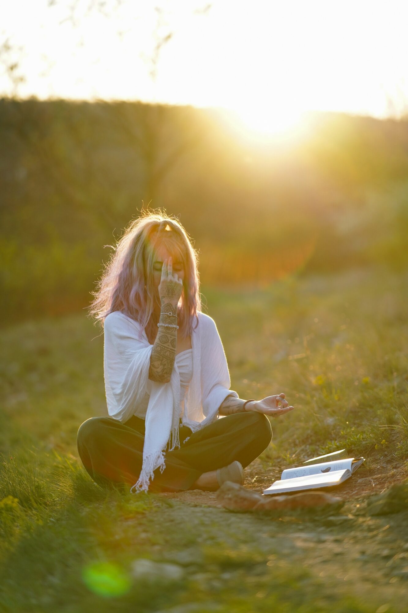 Person sitting cross-legged outdoors at sunset, holding a phone to their face, with a book nearby on the ground.