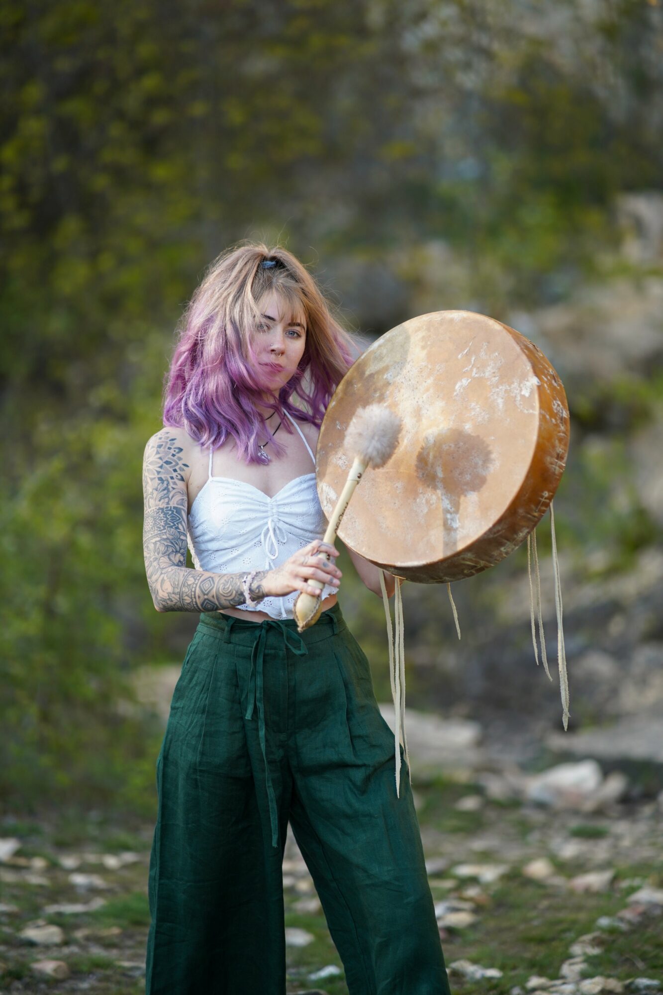 Young woman with purple hair holding a large drum and drumstick outdoors, trees and rocks in background.