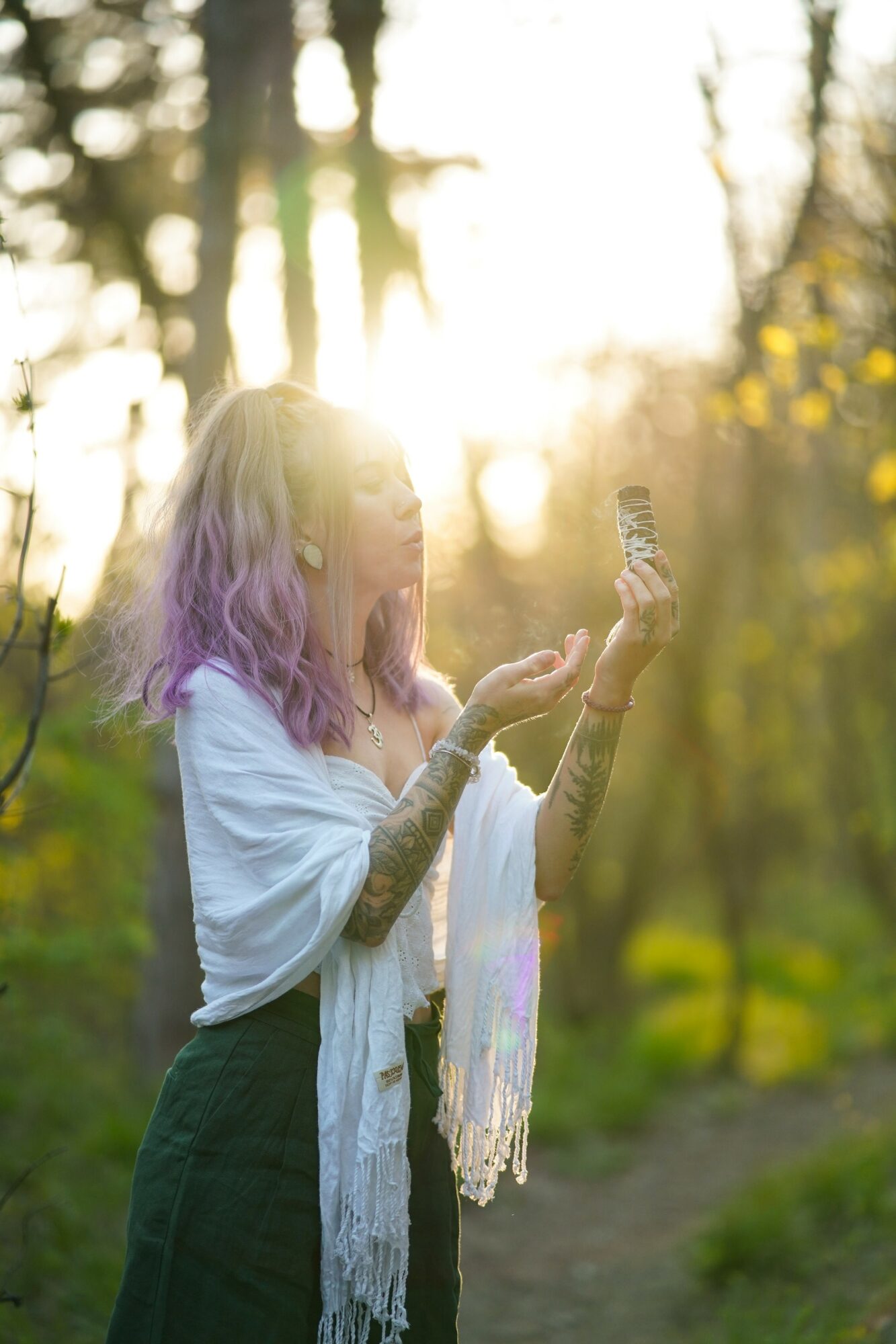 Young woman with purple hair outdoors, looking at her phone, sunlight filtering through trees, autumn scenery.