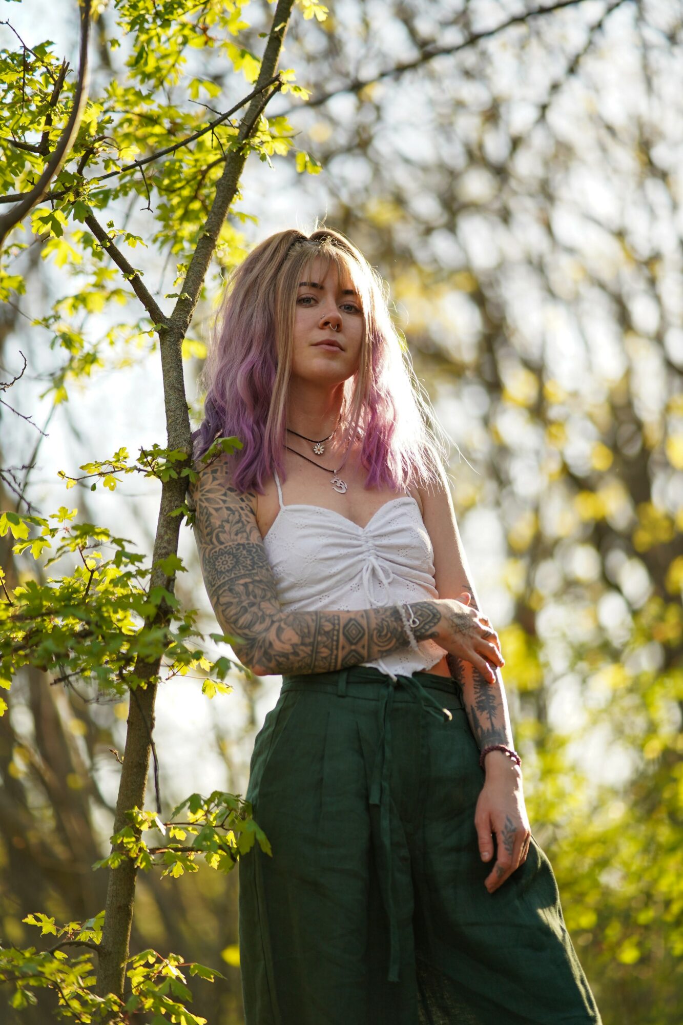 Young woman with purple hair and tattoos standing outdoors among green leaves, sunlight filtering through trees.