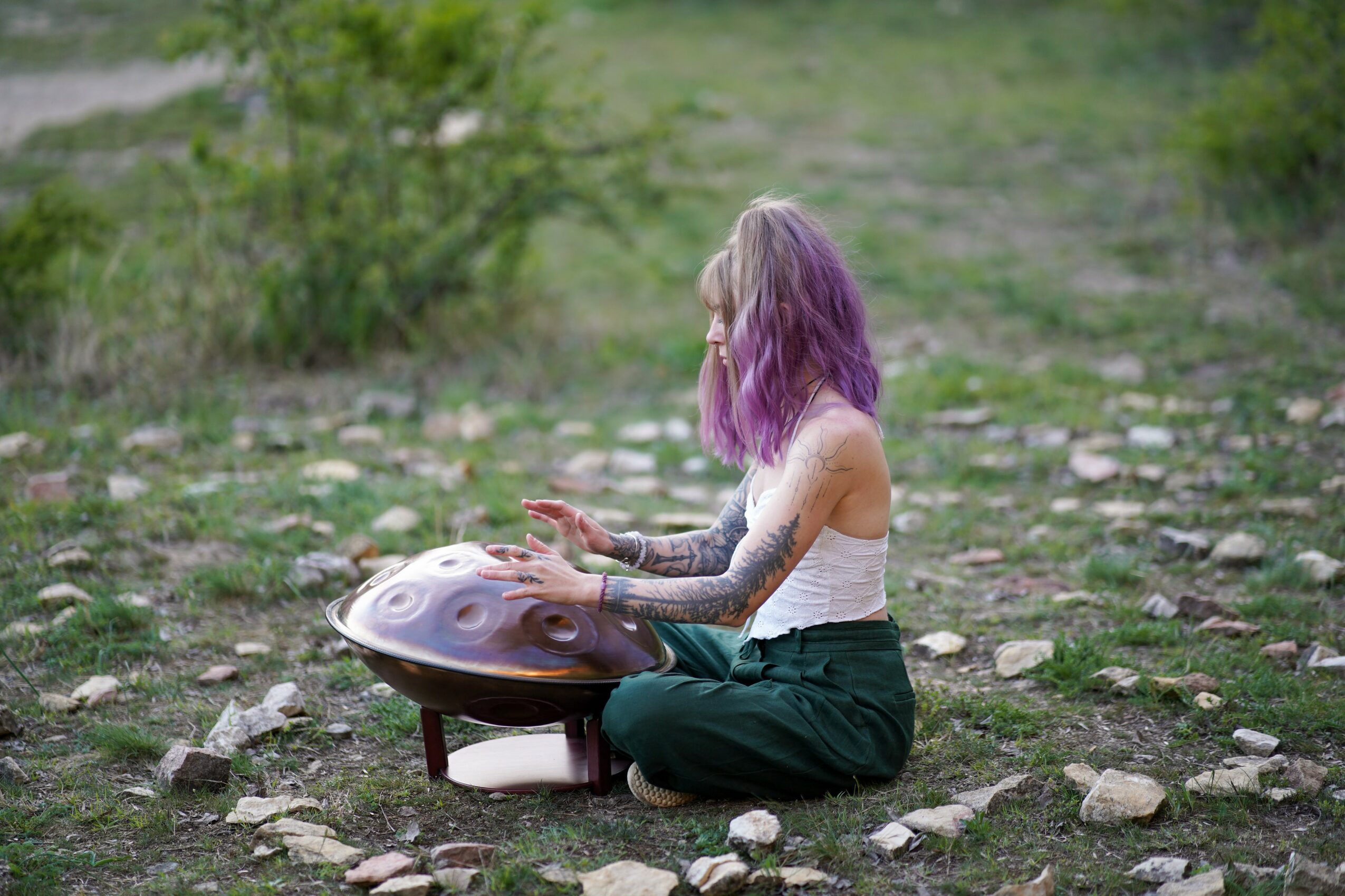 Person with purple hair sitting cross-legged outdoors, playing a handpan instrument on rocky ground.