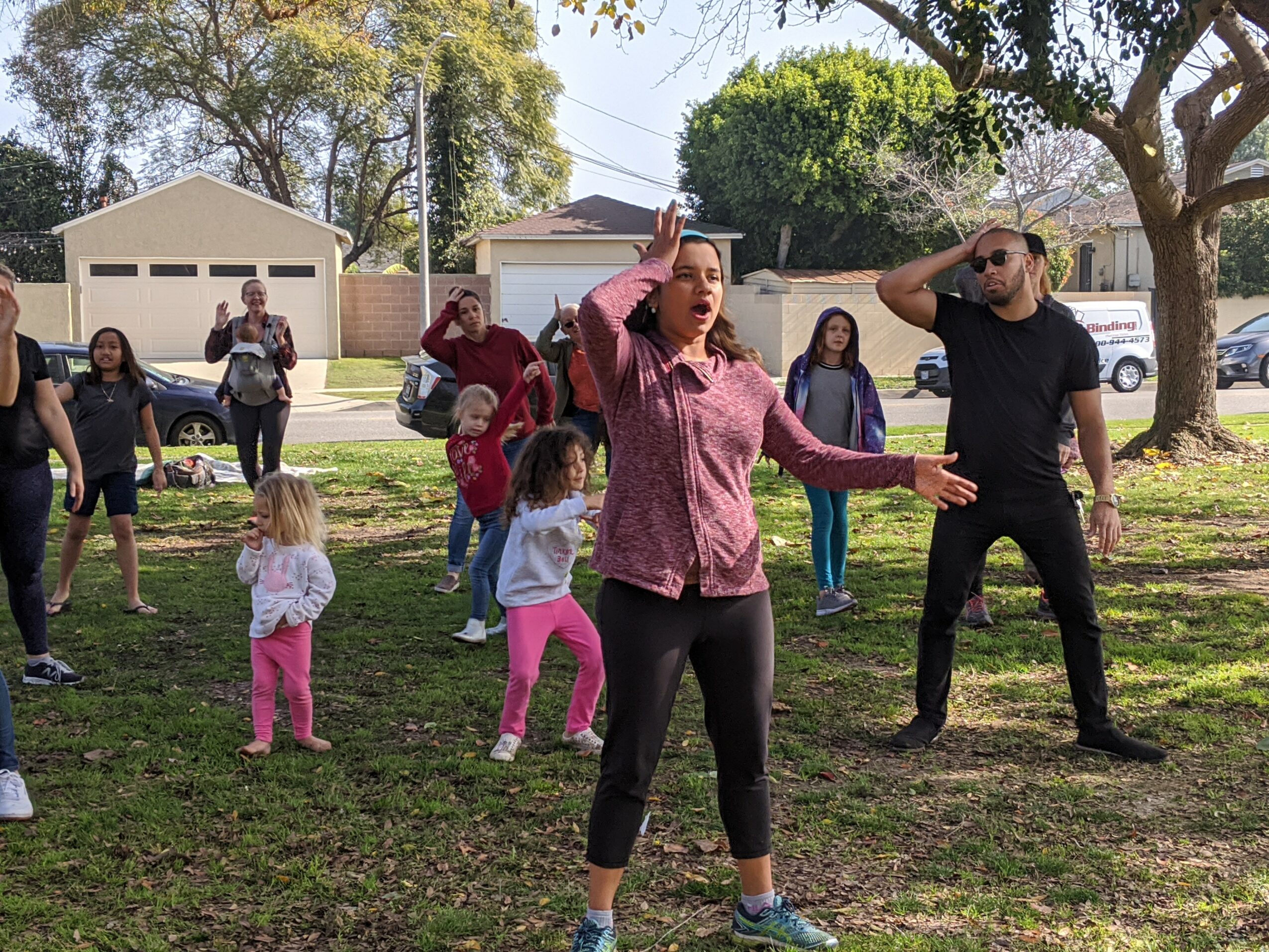 Group of people outdoors on grass, some children and adults, trees and houses in background, sunny day.
