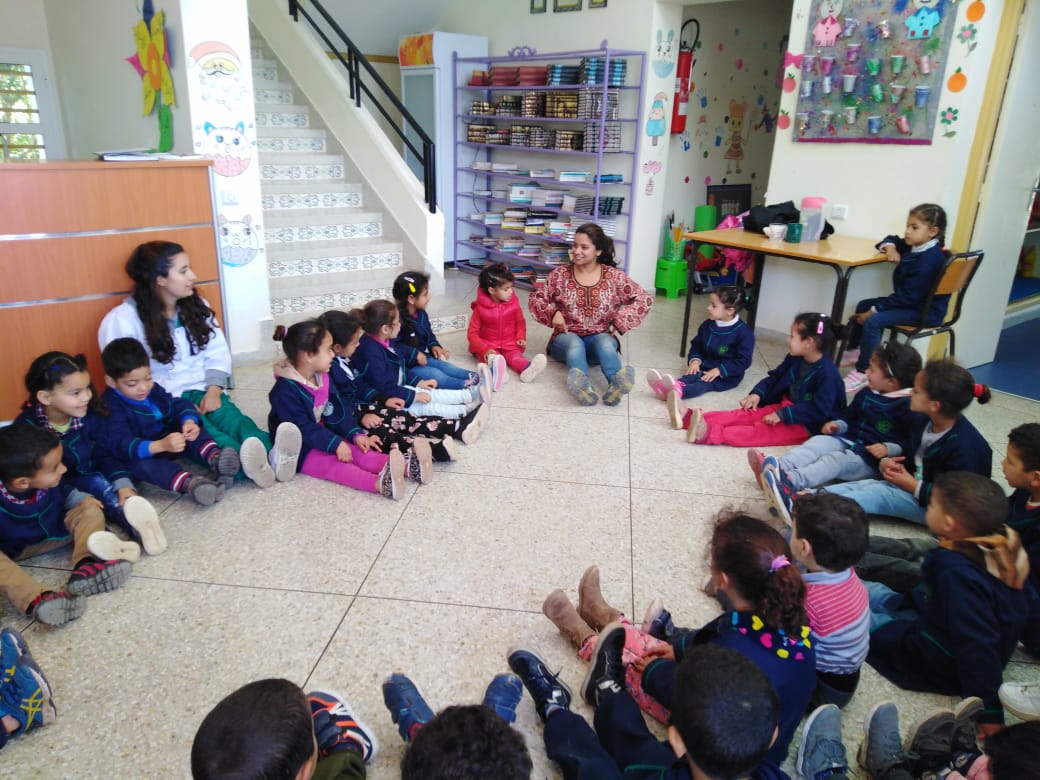 Teacher sitting on the floor with children sitting in a circle in a classroom, colorful decorations and stairs in background.