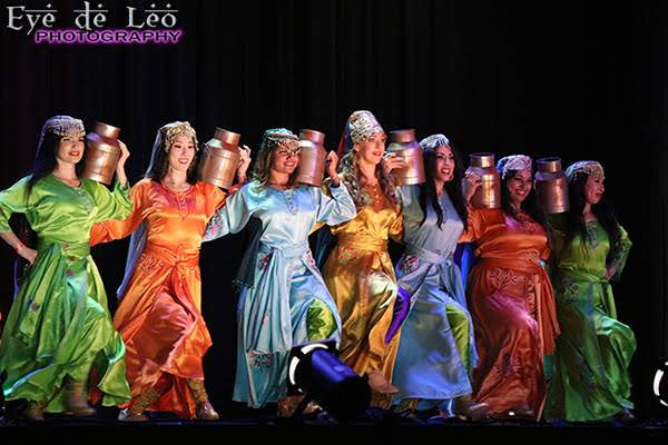 Group of women in colorful traditional costumes performing on stage, holding copper pots on their heads.