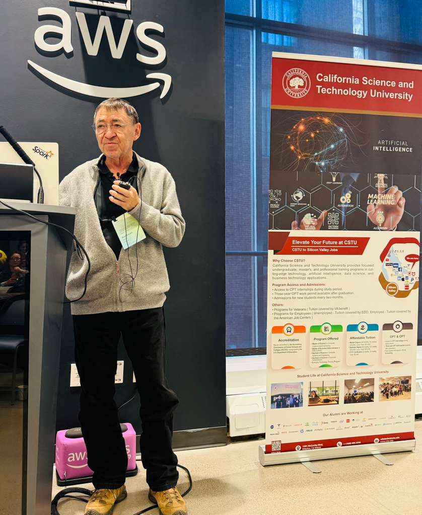Man speaking at a podium with AWS logo, standing next to a California Science and Technology University poster, in a conference setting.