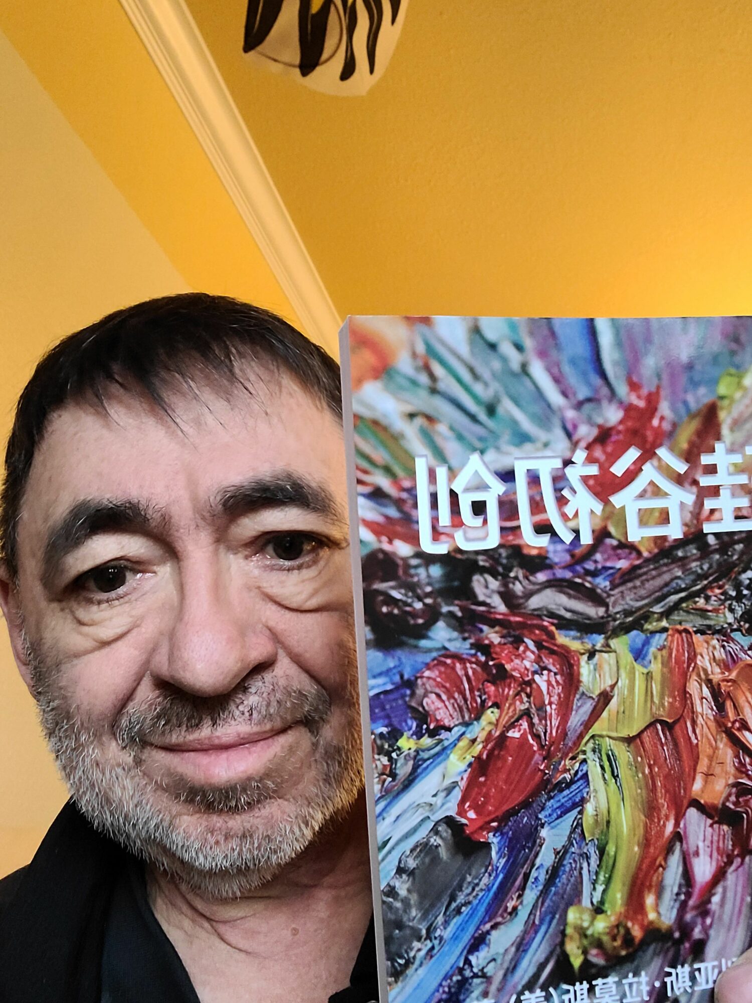 Close-up of a smiling man with short dark hair and a beard, against a yellow wall with a colorful abstract painting.
