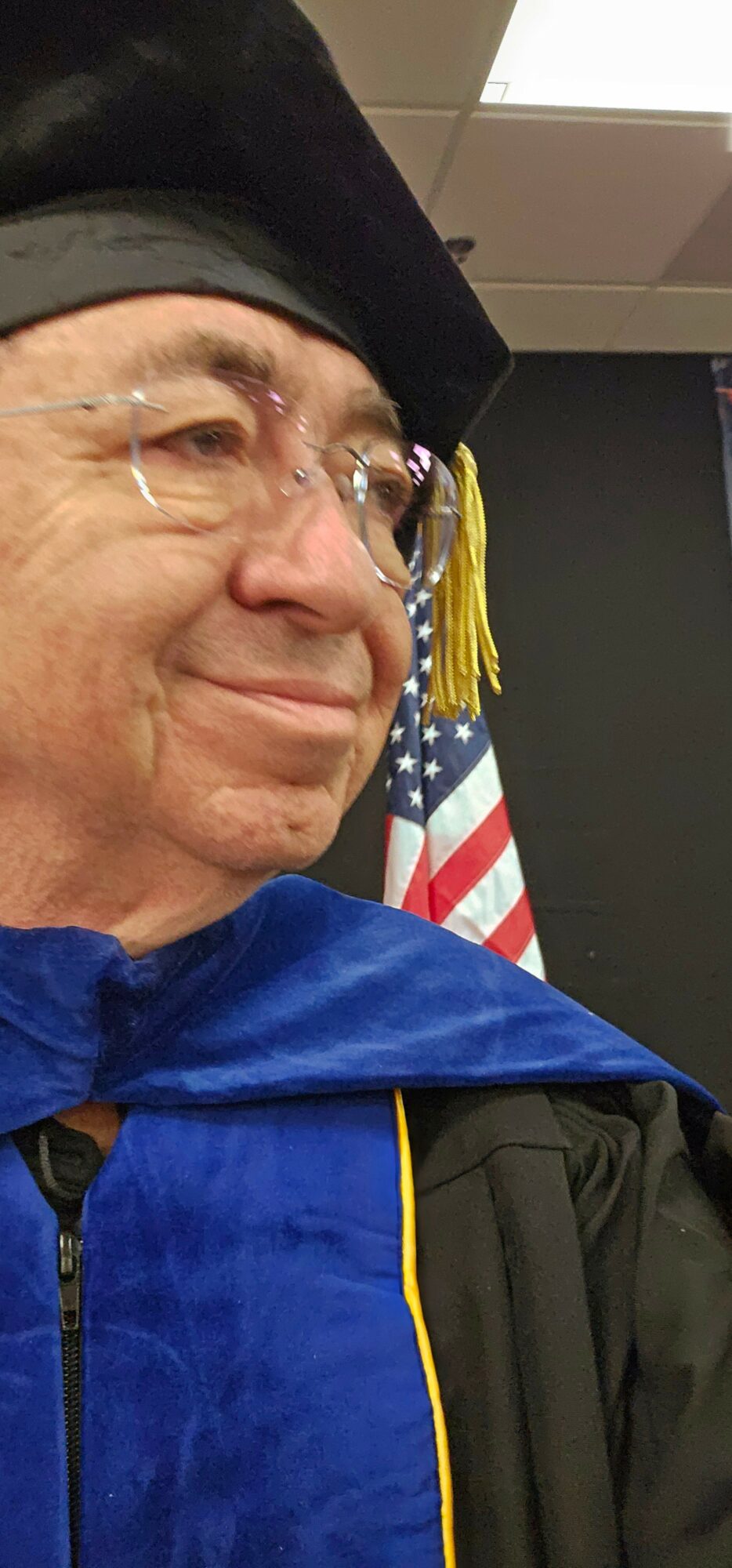 Person in graduation cap and gown, smiling, with American flag in background.