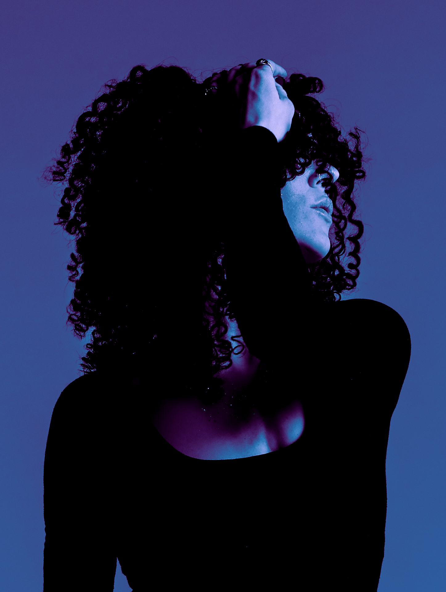 Person with curly hair touching their head, face turned sideways, illuminated with blue and purple lighting.