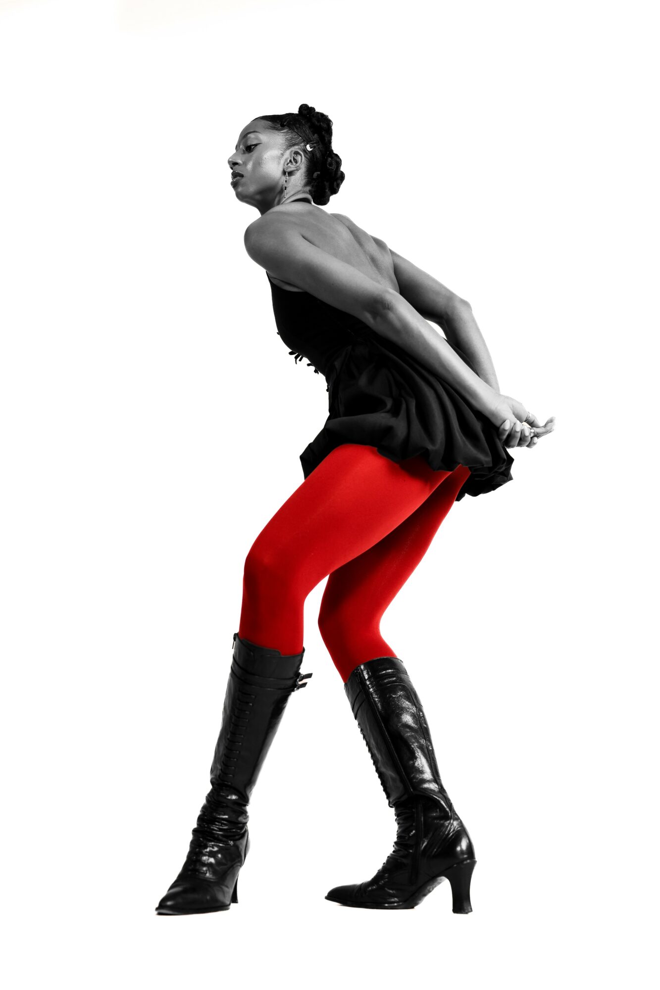 A woman in a dance pose with hands behind her back, wearing red tights and black boots, against a white background.