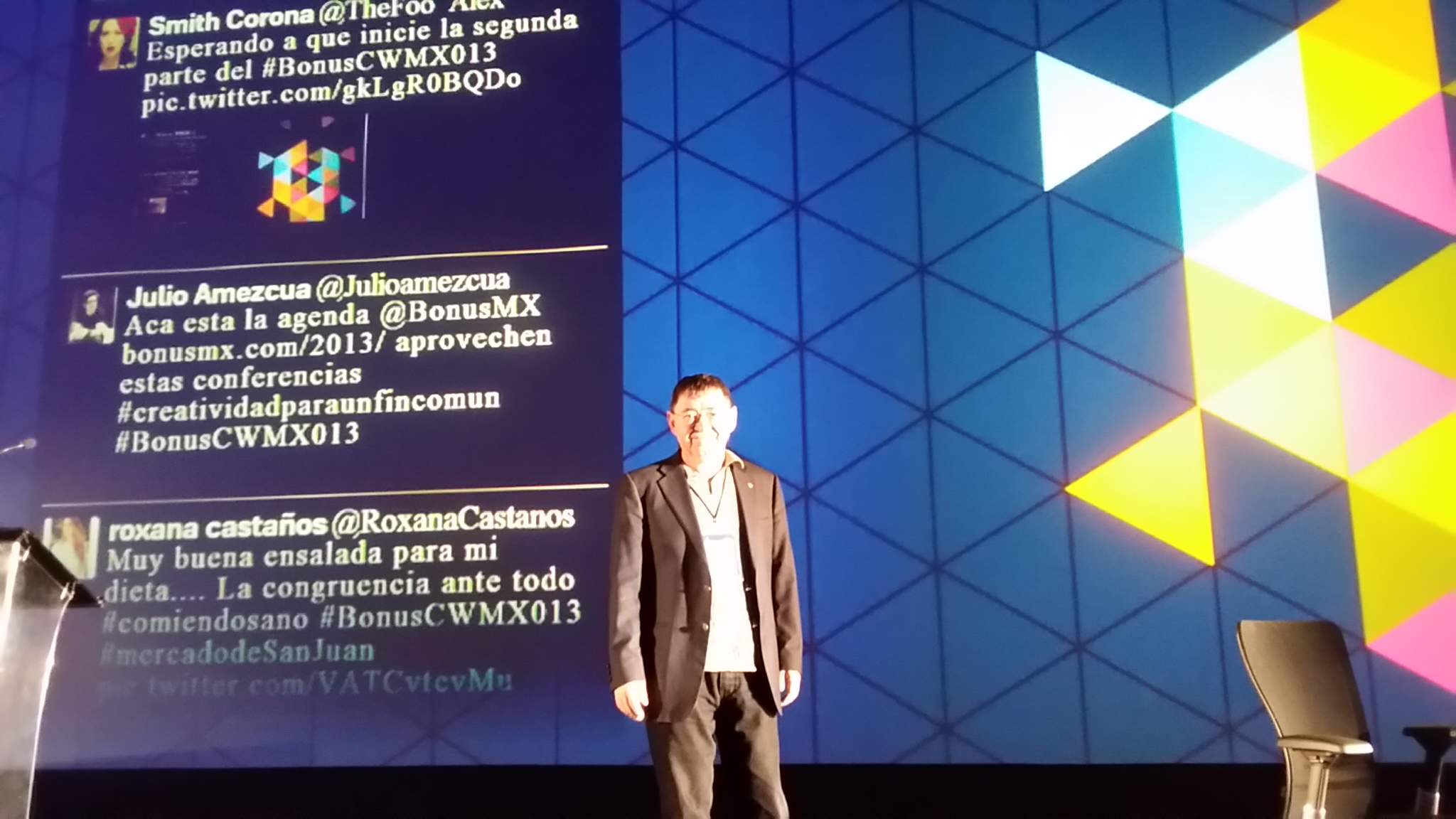 Man in suit standing on stage with a large colorful geometric background and text projected behind him.