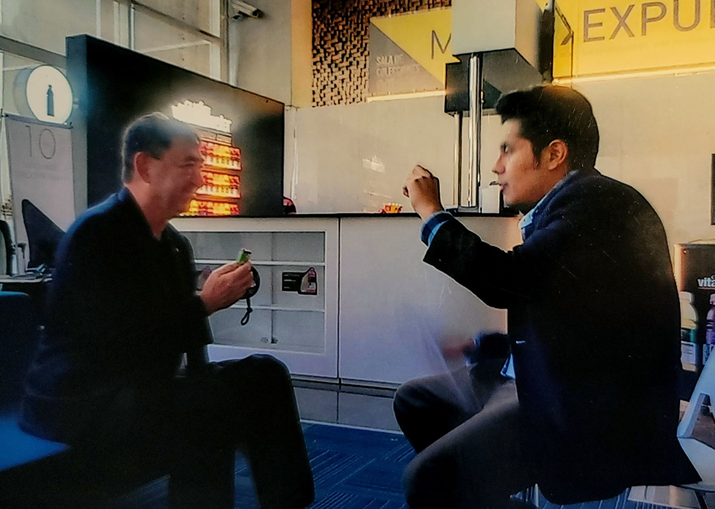 Two men are standing and talking in an indoor setting with a vending machine and a yellow sign in the background.