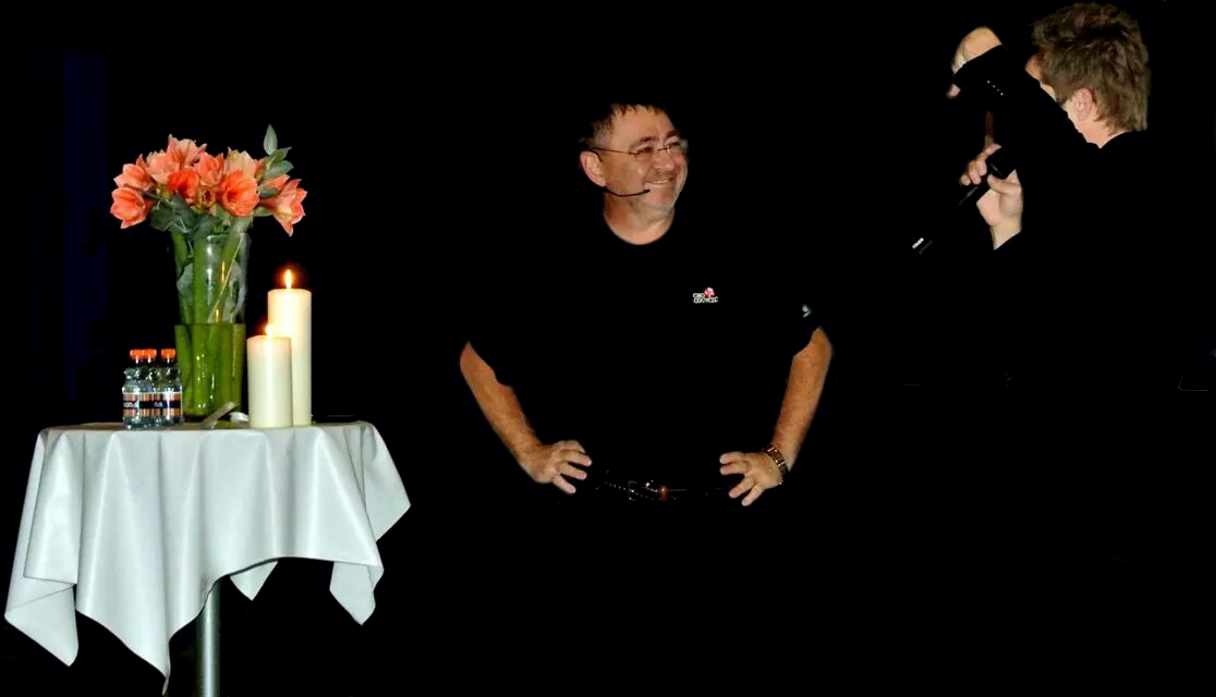Group of people talking, with a table holding flowers, candles, and drinks on the left side, against a dark background.