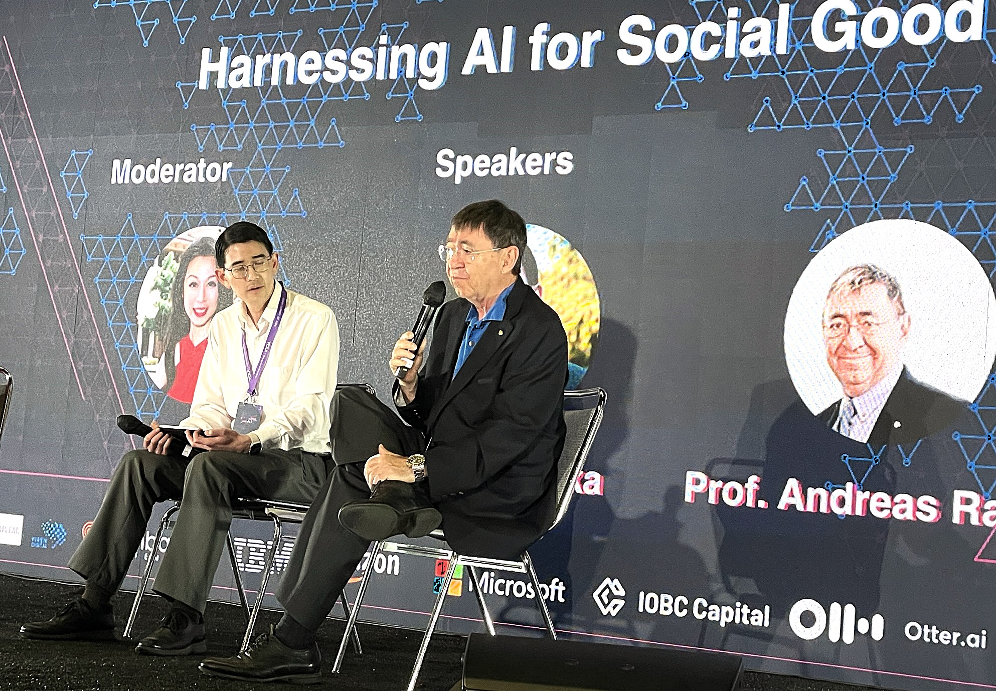 Two men seated on stage, one speaking into a microphone, with a large screen behind them displaying event information.