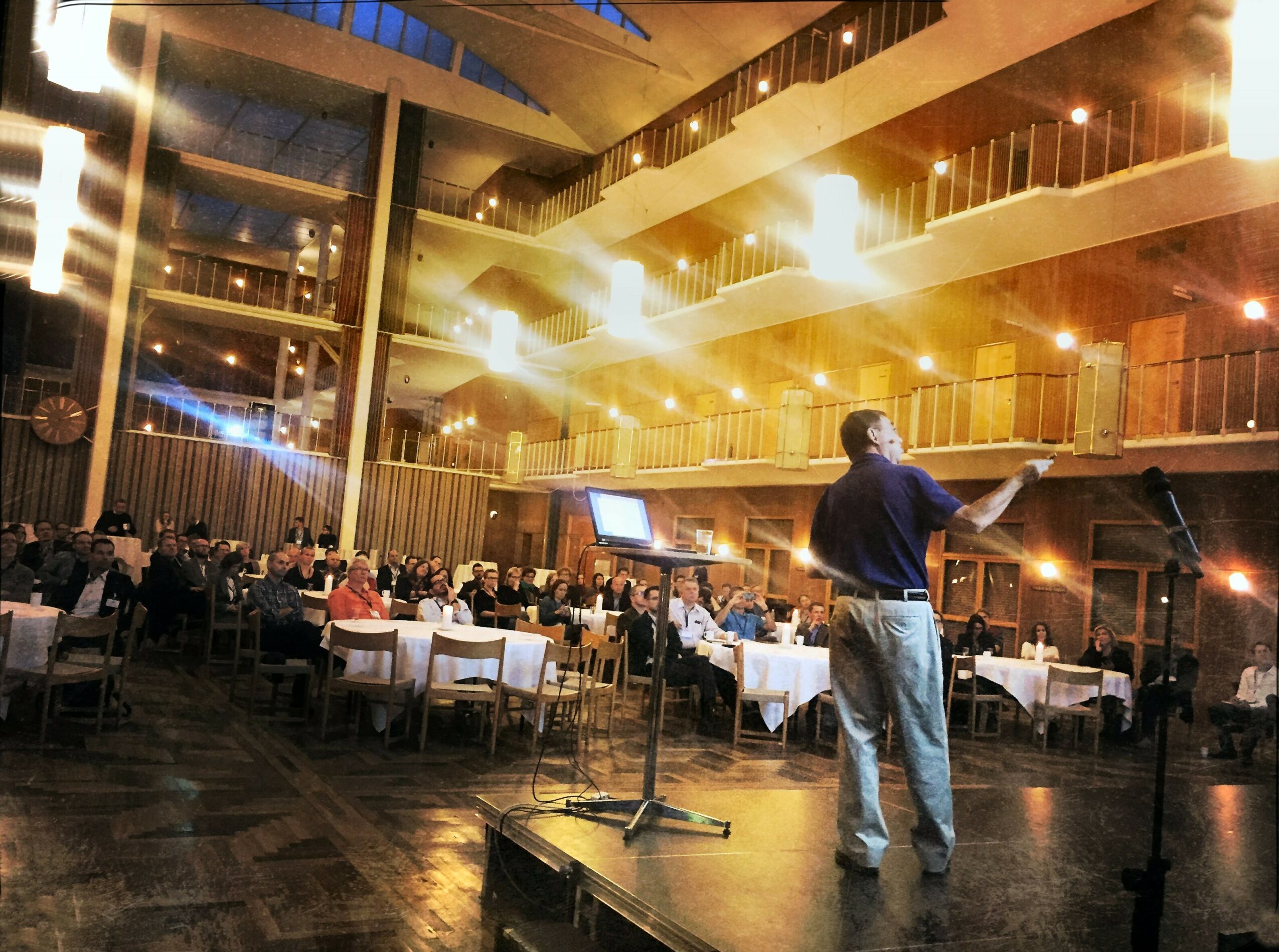 Person on stage speaking to an audience in a large, multi-level venue with warm lighting and tables.