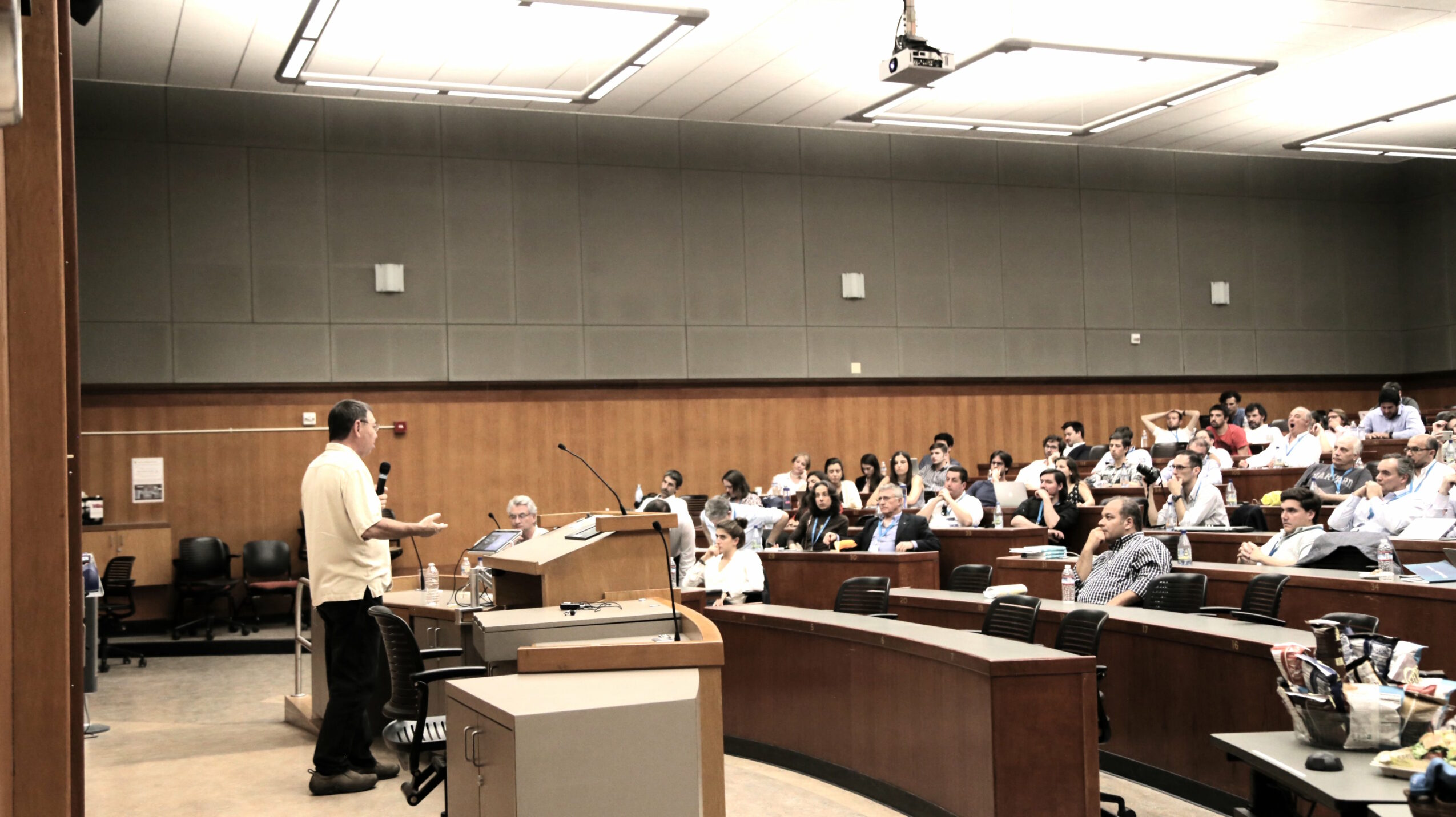 Lecture hall with a speaker at a podium and audience seated in tiered rows.