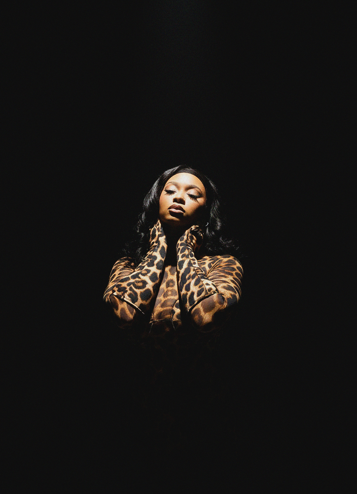 Woman with leopard print clothing and dark hair poses with hands near face against black background.