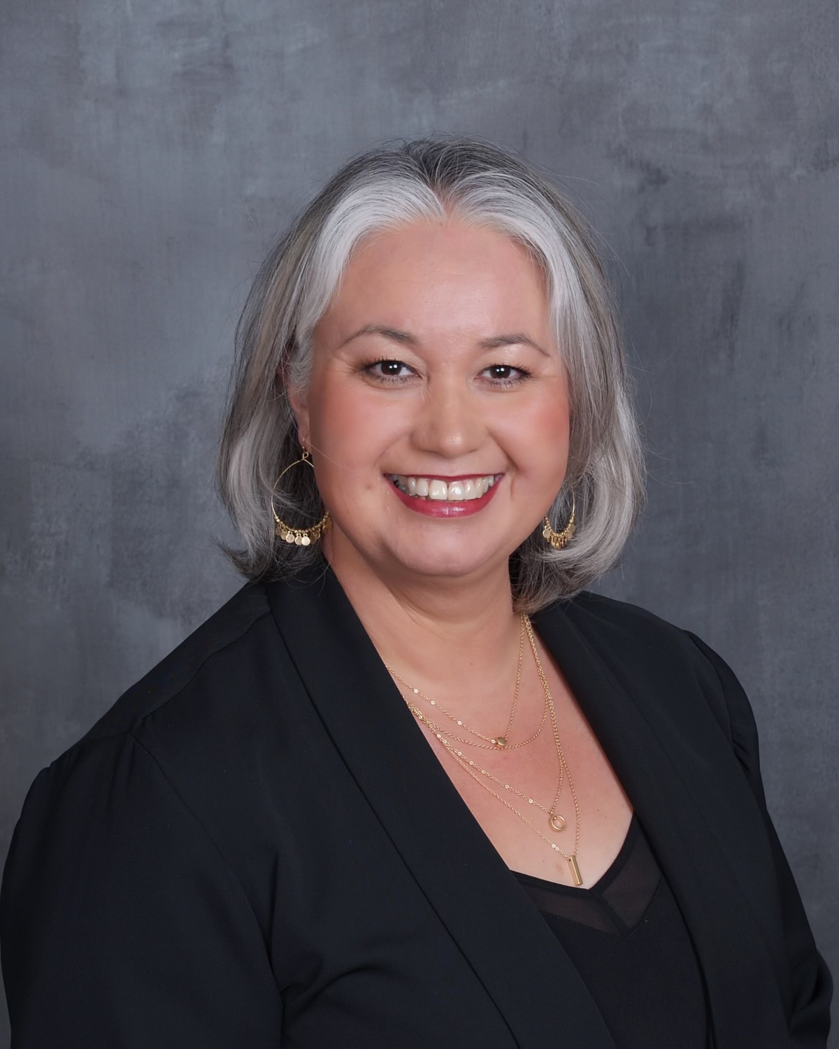 Smiling woman with shoulder-length gray hair, wearing a black blazer and gold jewelry, against a gray background.