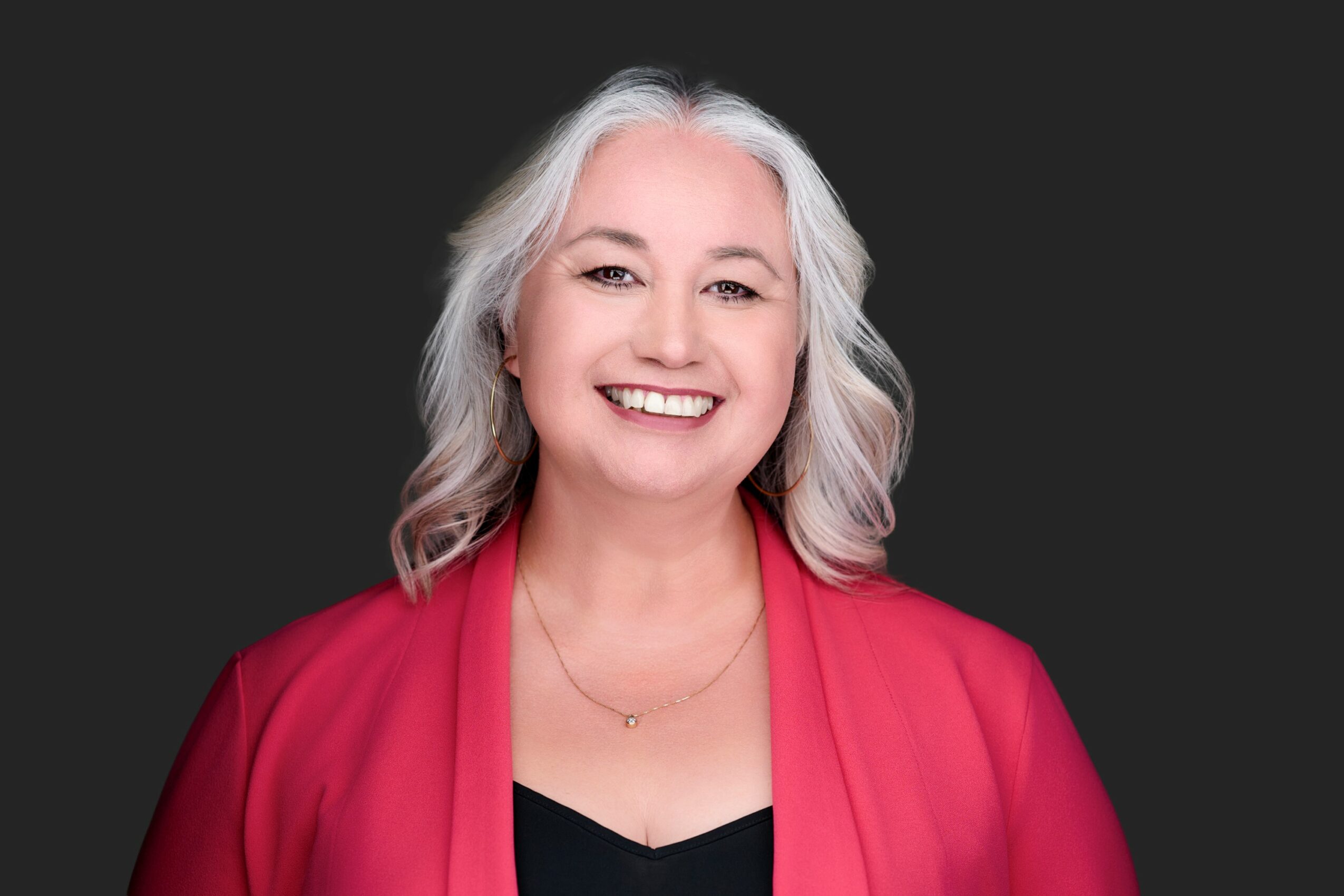 Smiling woman with shoulder-length gray hair wearing a red blazer and black top, against a dark background.