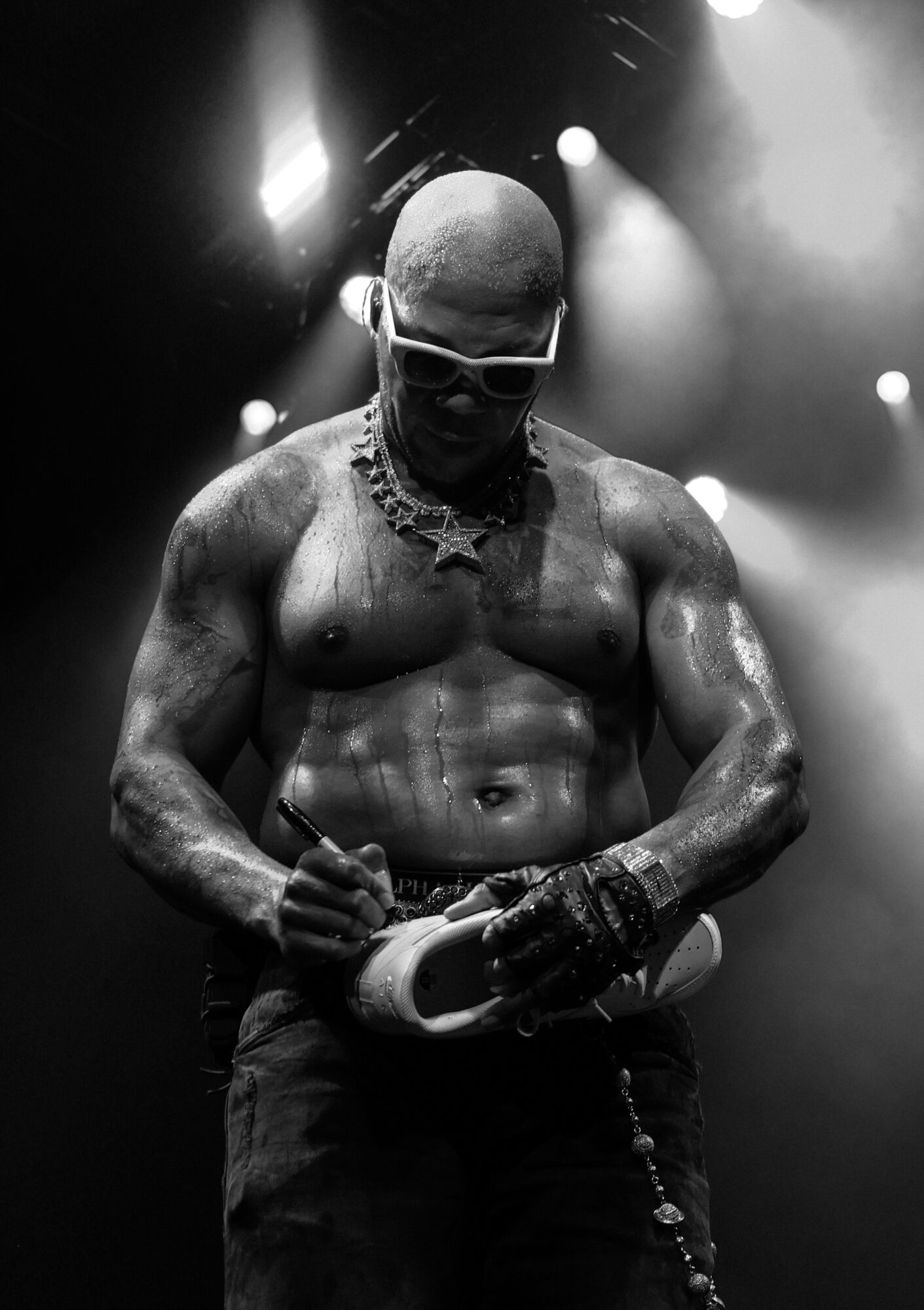 Muscular man with sunglasses and jewelry, holding a microphone, on stage with bright lights behind him.