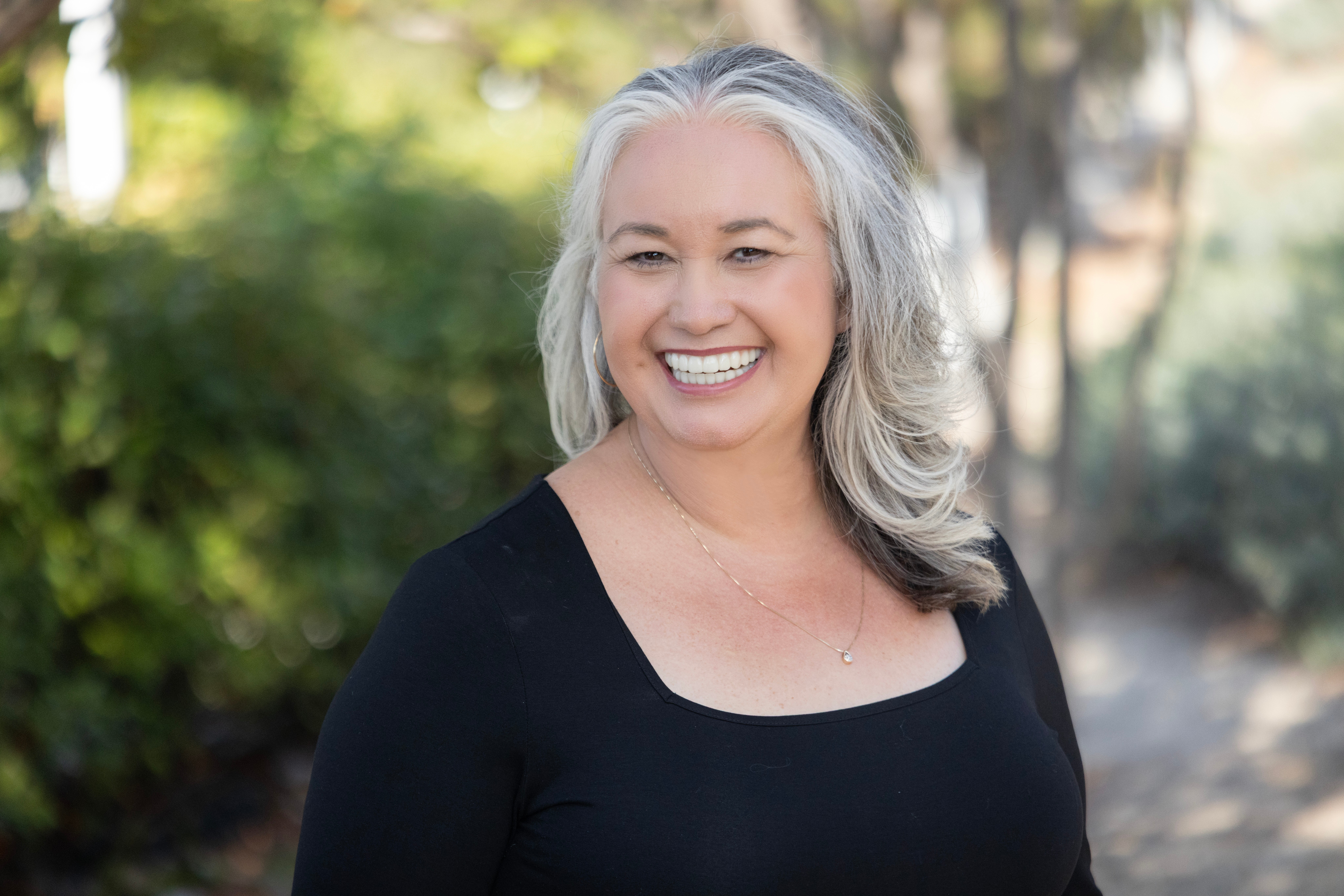 Smiling woman with gray hair outdoors, wearing a black top, surrounded by greenery.