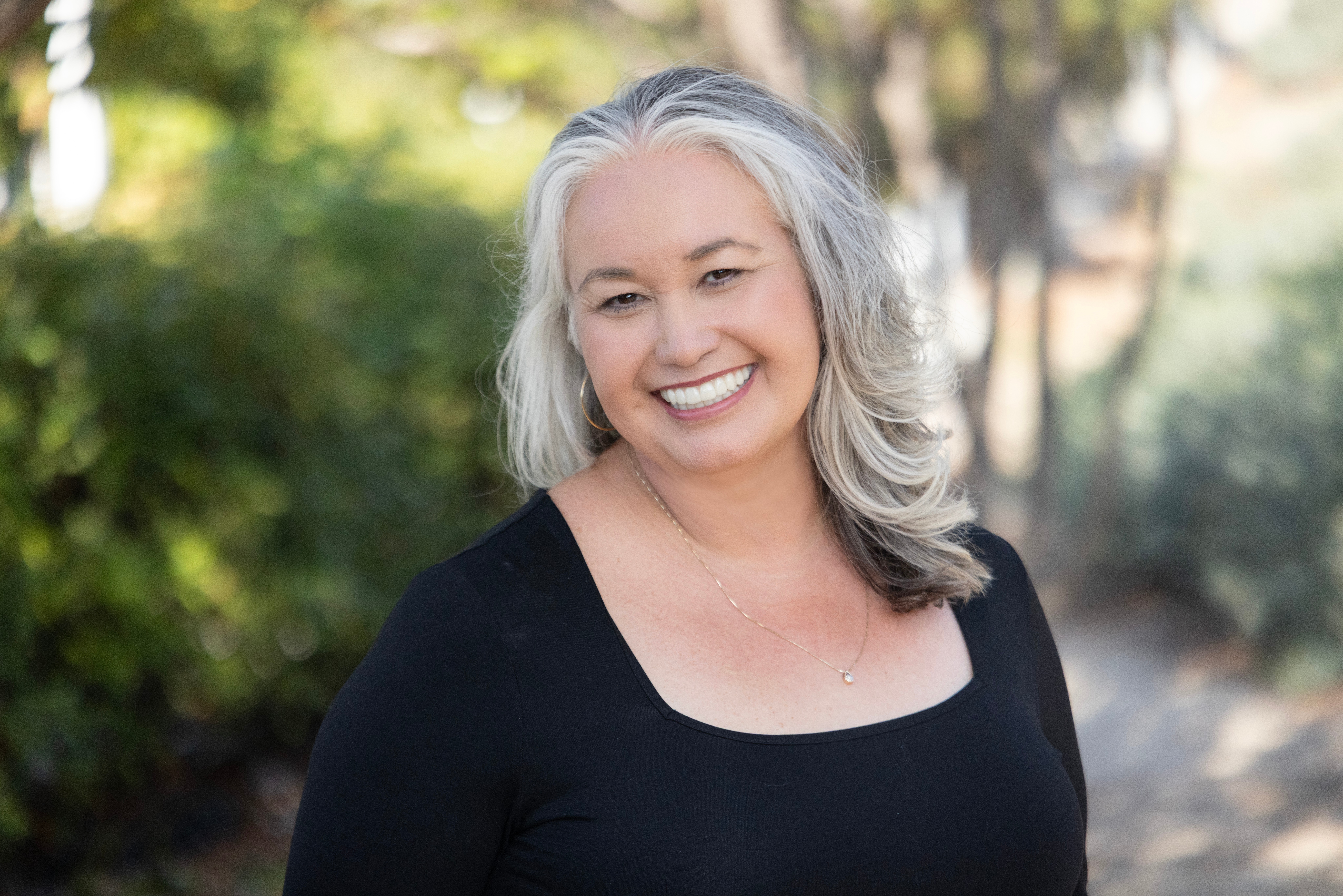 Smiling woman with gray hair outdoors, wearing a black top, surrounded by greenery.