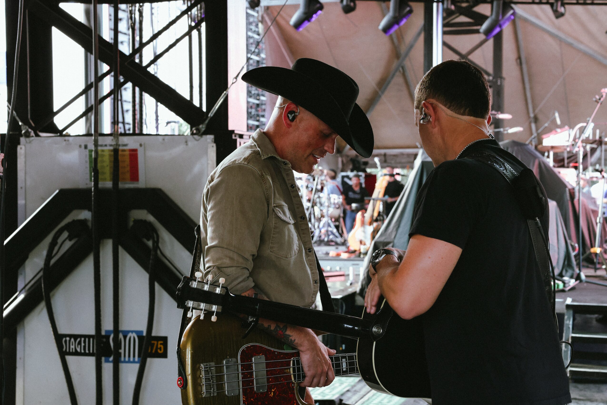 Two people on stage, one with a guitar and a wide-brimmed hat, the other with short hair, in a concert setting.