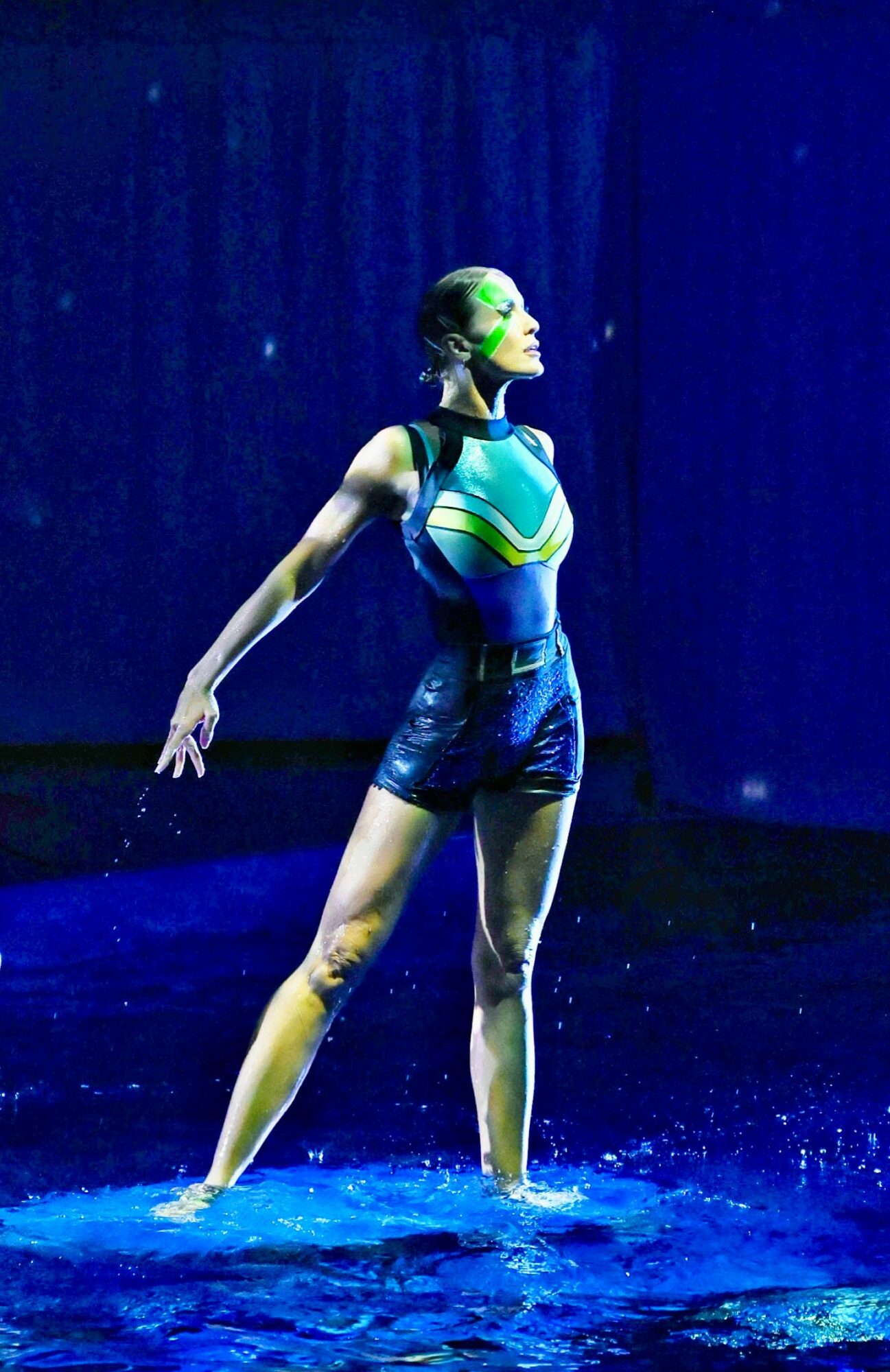 A woman stands in water with arms outstretched, wearing a colorful swimsuit and goggles, against a dark background.