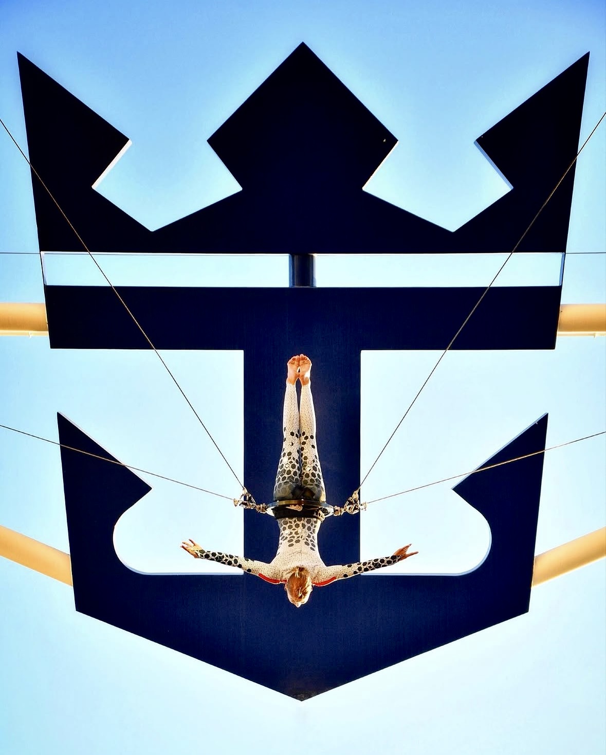 Person hanging upside down with arms outstretched, reflected in a mirror with a crown-shaped design, against a blue sky.
