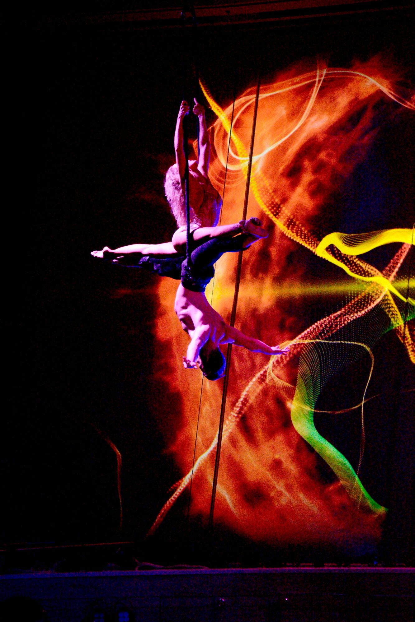 Two performers in costumes dance against a fiery background with colorful light trails, one upside down and one upright.