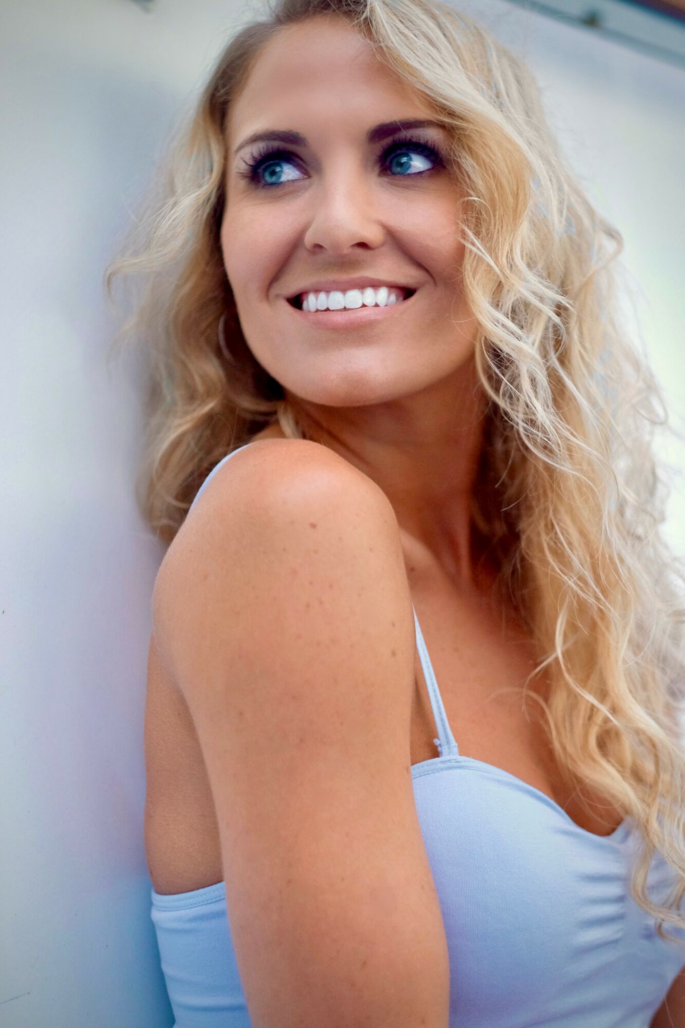 Smiling woman with long, curly blonde hair and blue eyes, wearing a light blue top, looking to the side.