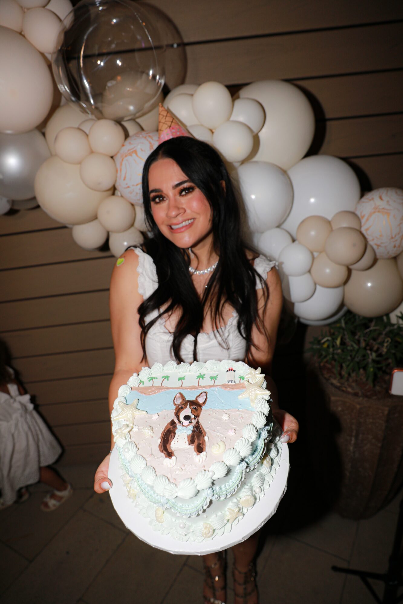 Young woman with long dark hair holding a decorated cake with a dog illustration, standing in front of balloon decorations.