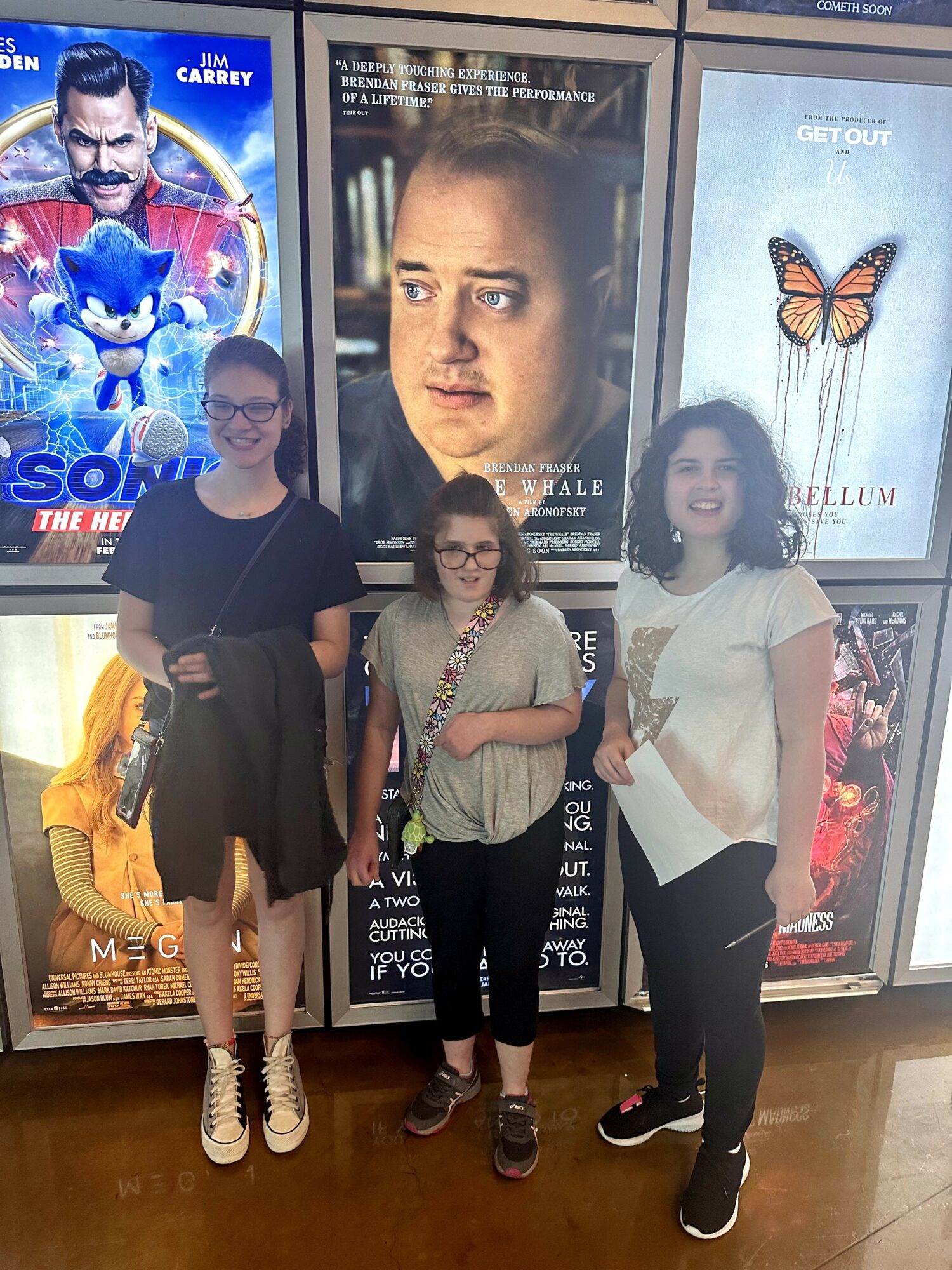 Three girls stand in front of movie posters at a theater, smiling and posing for the photo.