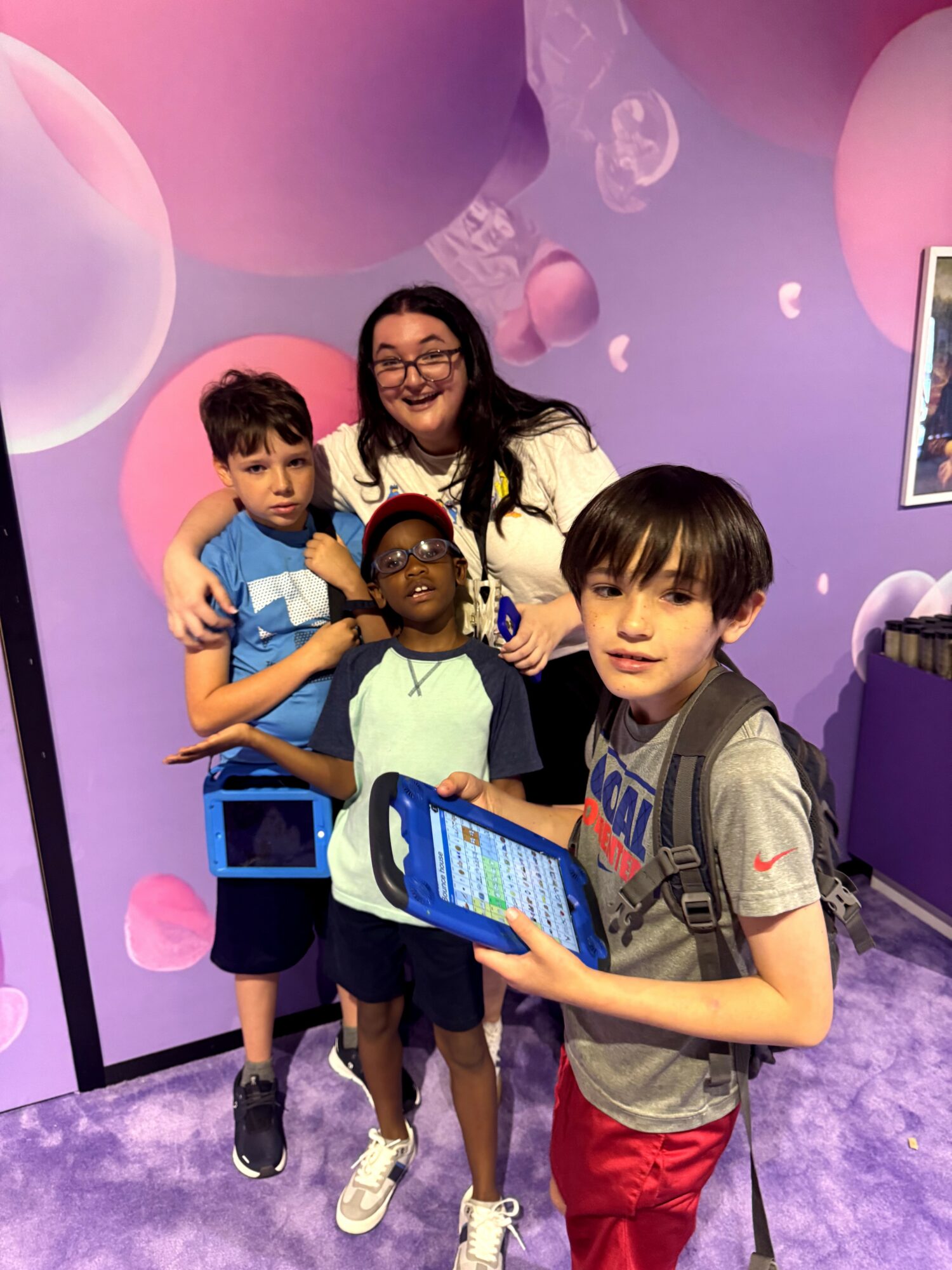 Group of four children and a woman smiling, with colorful circular background, one child holding a tablet, in an indoor setting.
