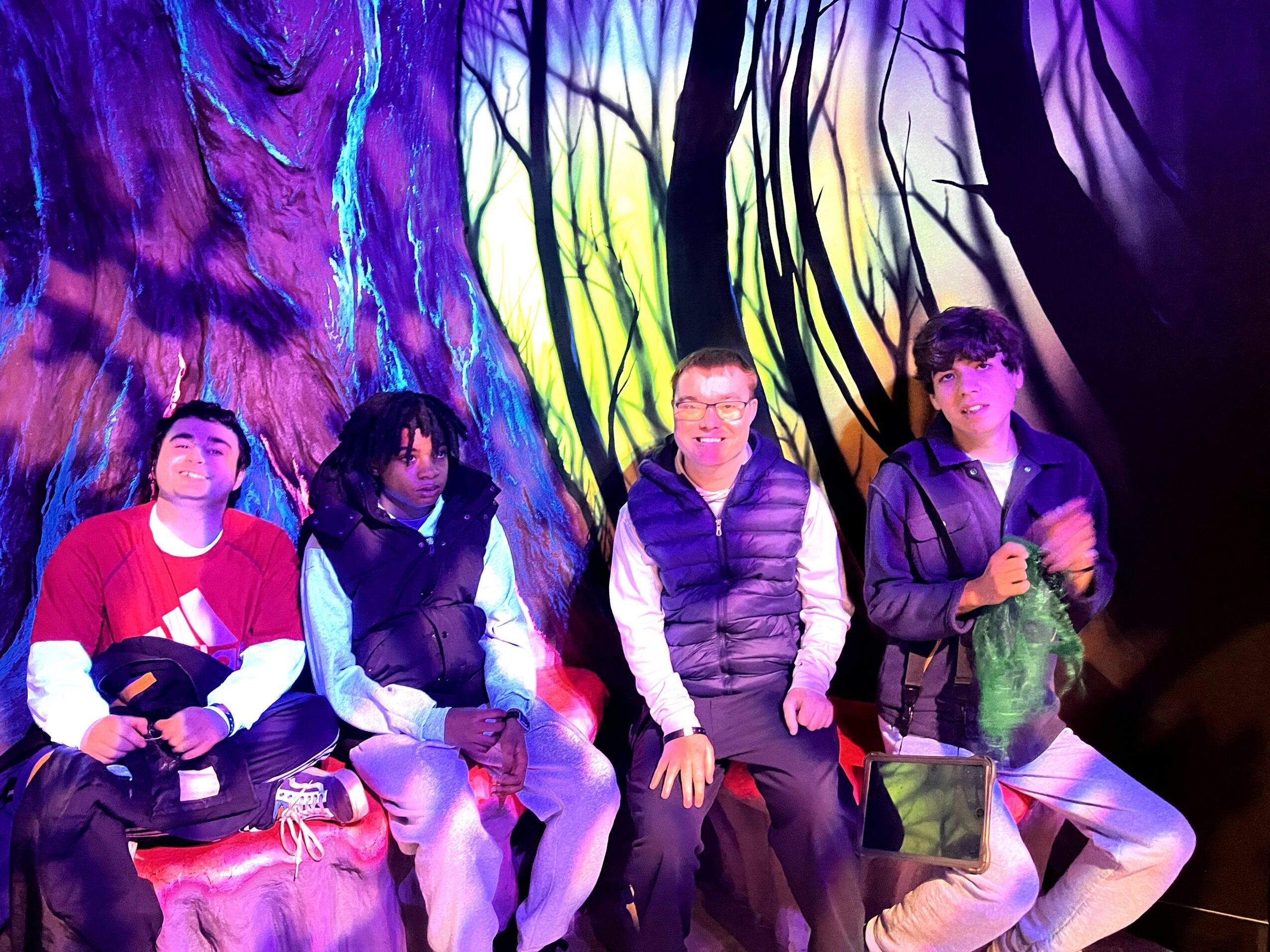 Four people sitting in front of a colorful forest backdrop, some holding objects, with vibrant lighting.